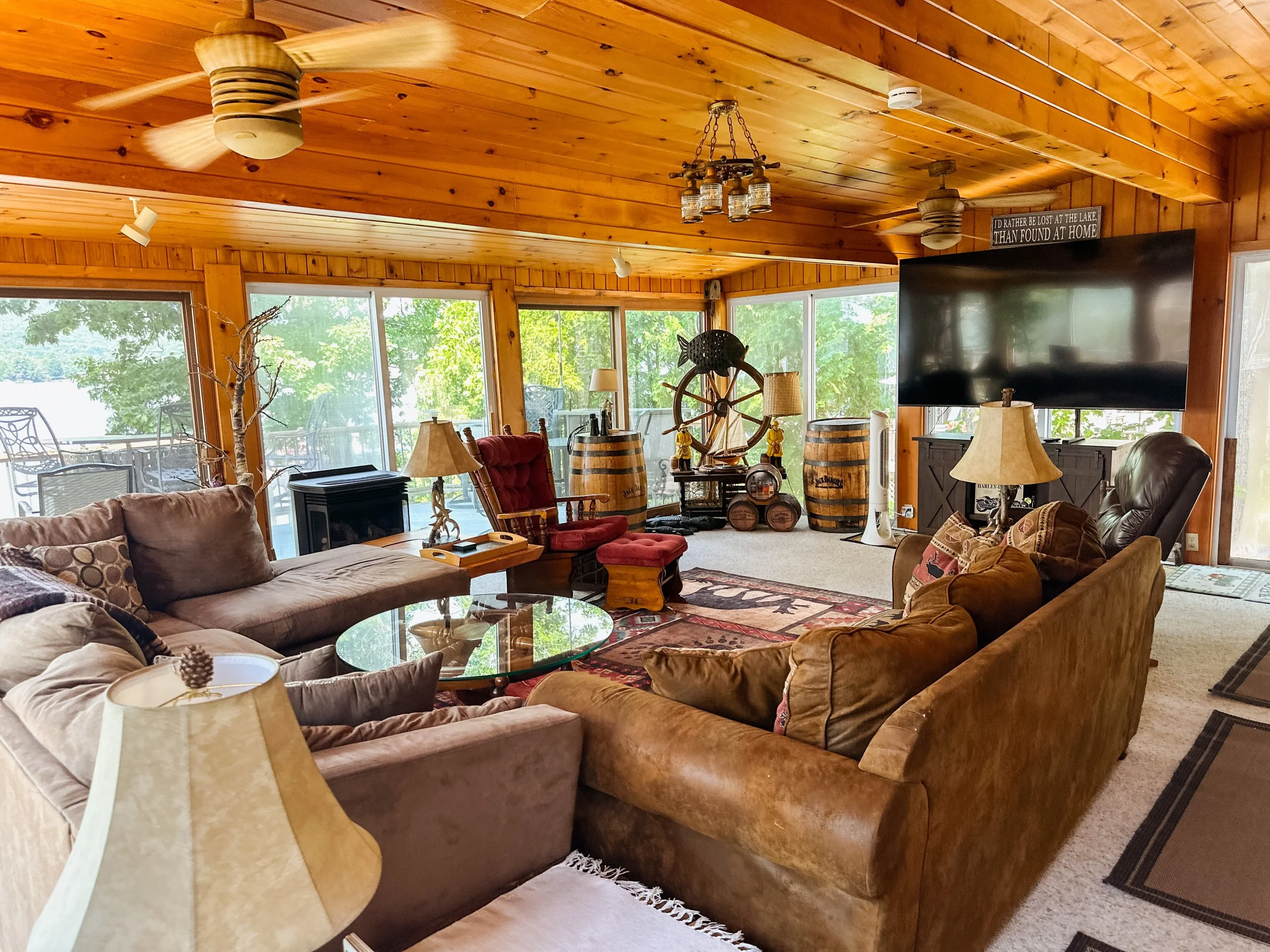 A cozy living room with wooden walls and ceiling, filled with furniture including sofas, chairs, and lamps. Large windows reveal green trees outside. Decorative items like a ship wheel and barrels are present, and a television is mounted on the wall.