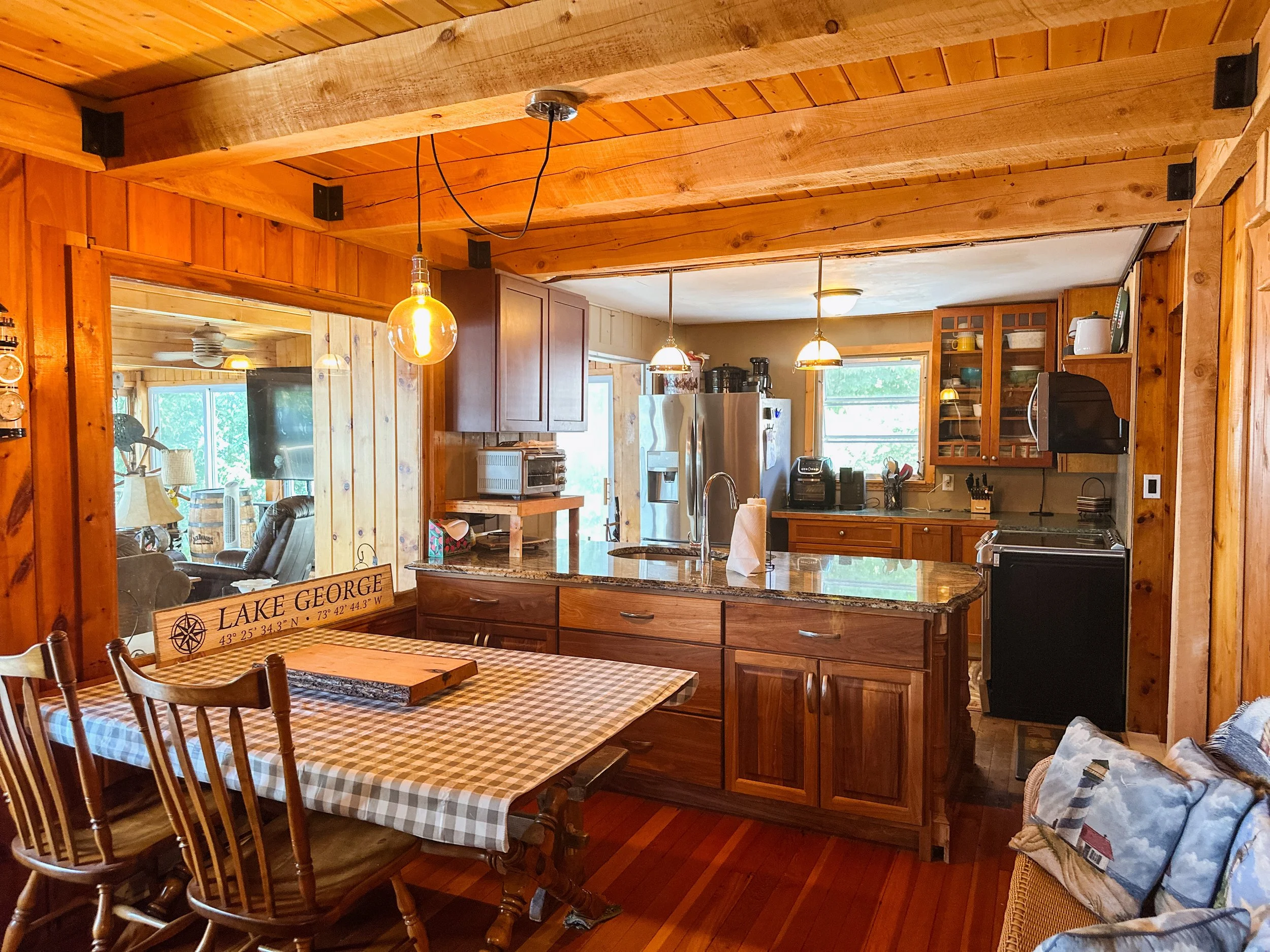 View of a cozy kitchen and dining area with wooden ceiling, cabinets, and dining table with checkered tablecloth.