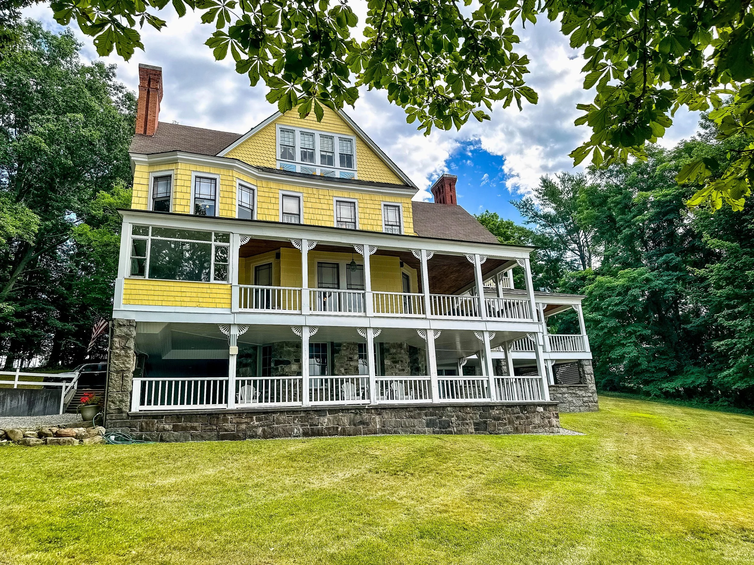 A yellow house with a porch, multiple windows, a chimney, surrounded by lush green trees and a well-maintained lawn under a partly cloudy sky.