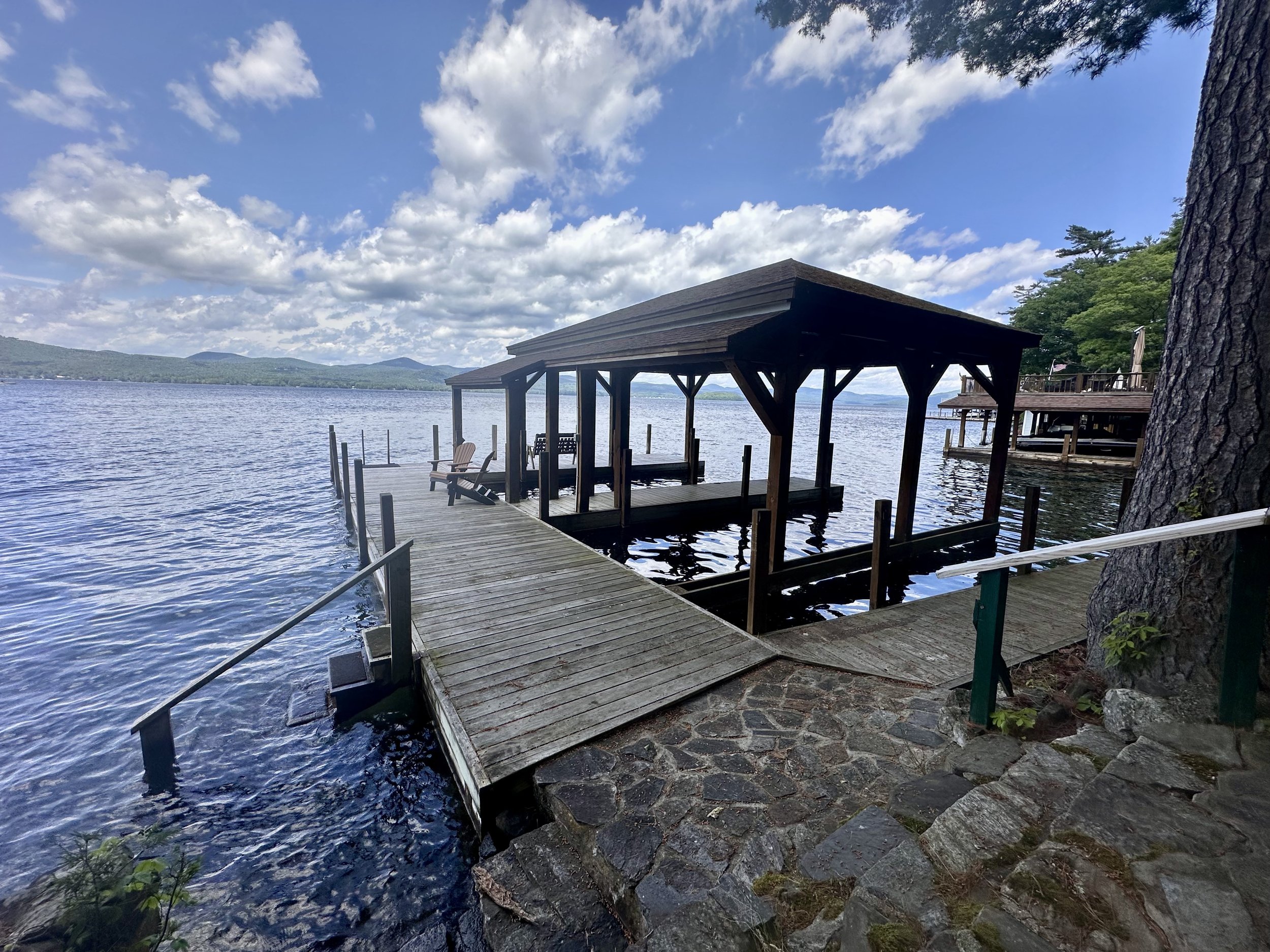 A wooden dock with a covered boat slip extending into a calm lake, with Adirondack chairs inside the slip, surrounded by trees and a partly cloudy sky.