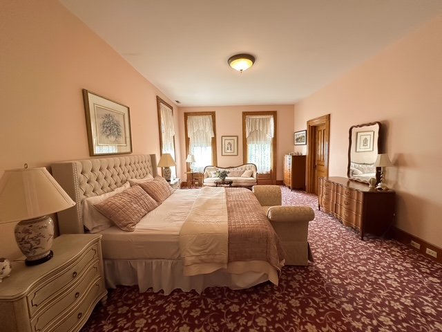 Spacious bedroom with a large bed, twin windows with lace curtains, and vintage furniture including dressers and a cushioned armchair.