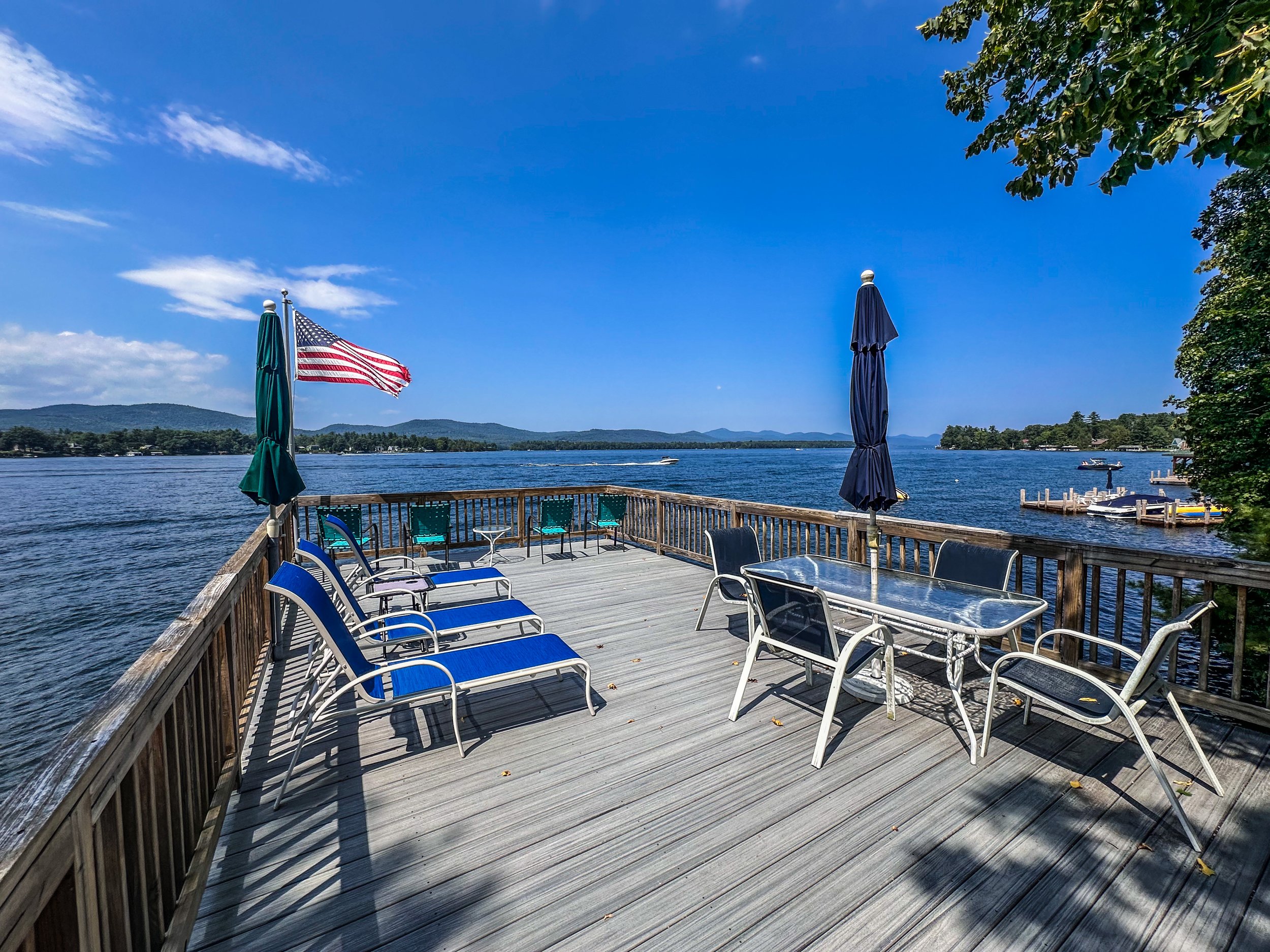 A waterfront patio with blue lounge chairs, a table, and umbrellas overlooking a river with boats and distant mountains under a partly cloudy sky.