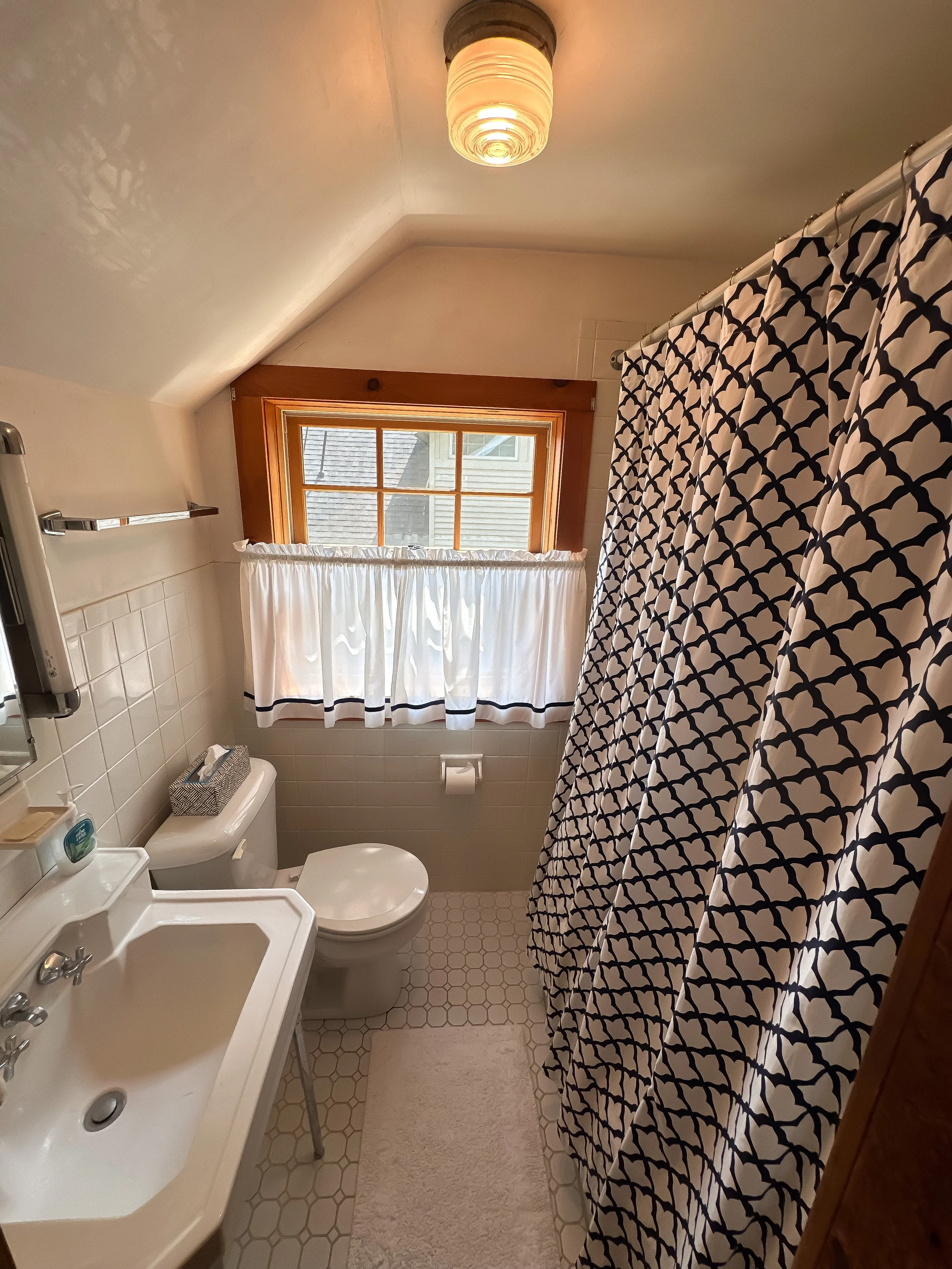 A small bathroom with a window covered by a white curtain, a patterned shower curtain, a white sink, and a toilet, illuminated by a ceiling light.