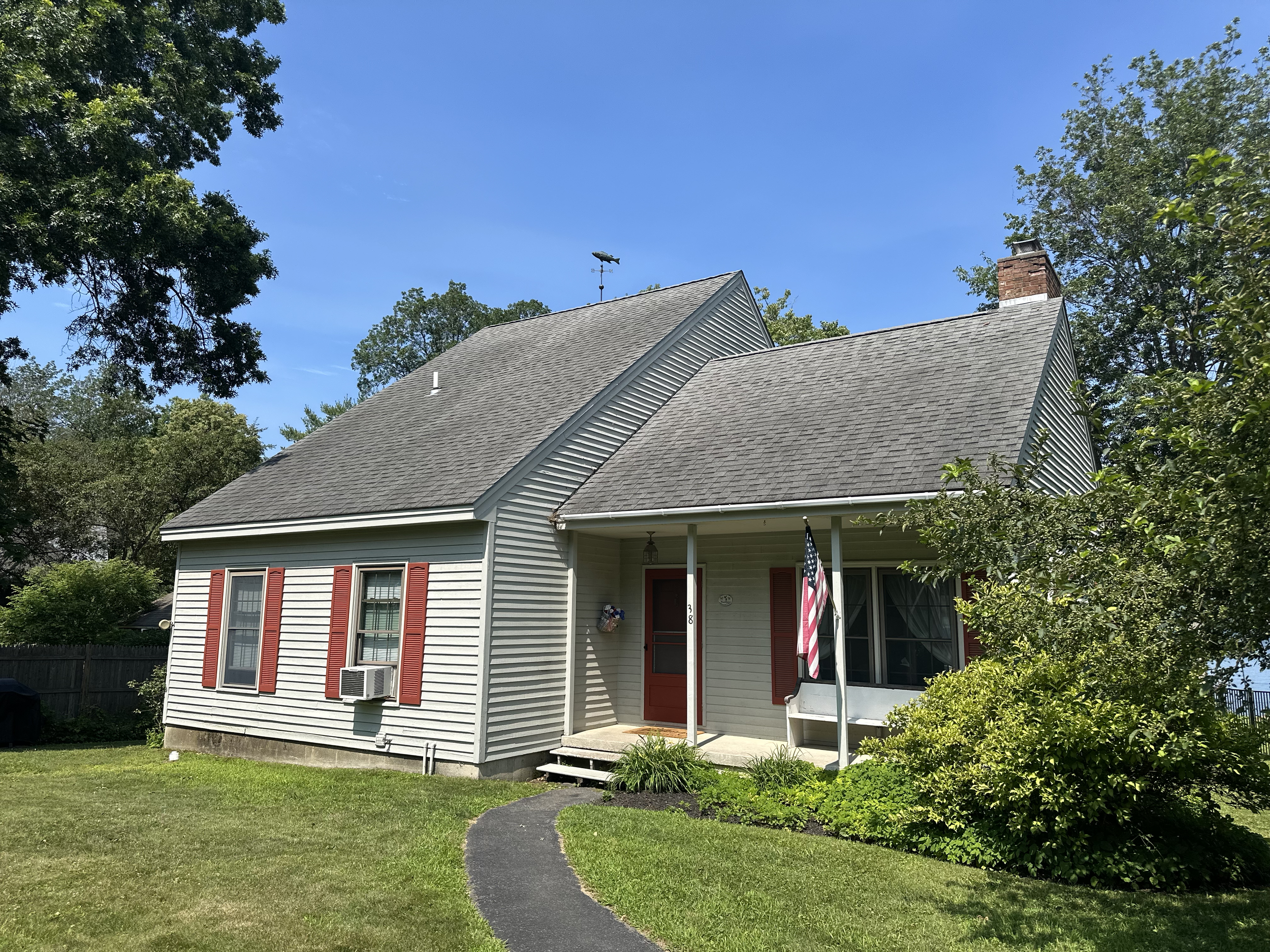 A white house with red shutters and a front porch, surrounded by green trees and grass, under a clear blue sky.