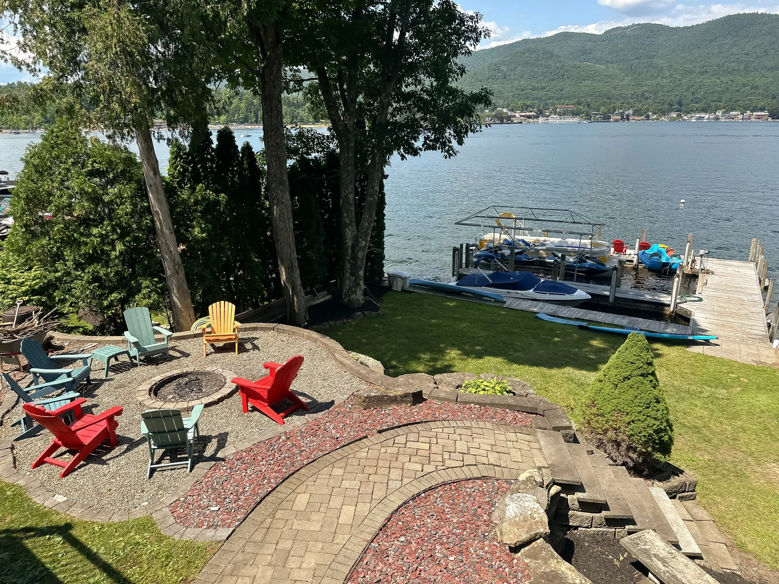 A lakeside backyard with colorful Adirondack chairs arranged around a fire pit, overlooking a body of water with boats and a distant town with mountains in the background.