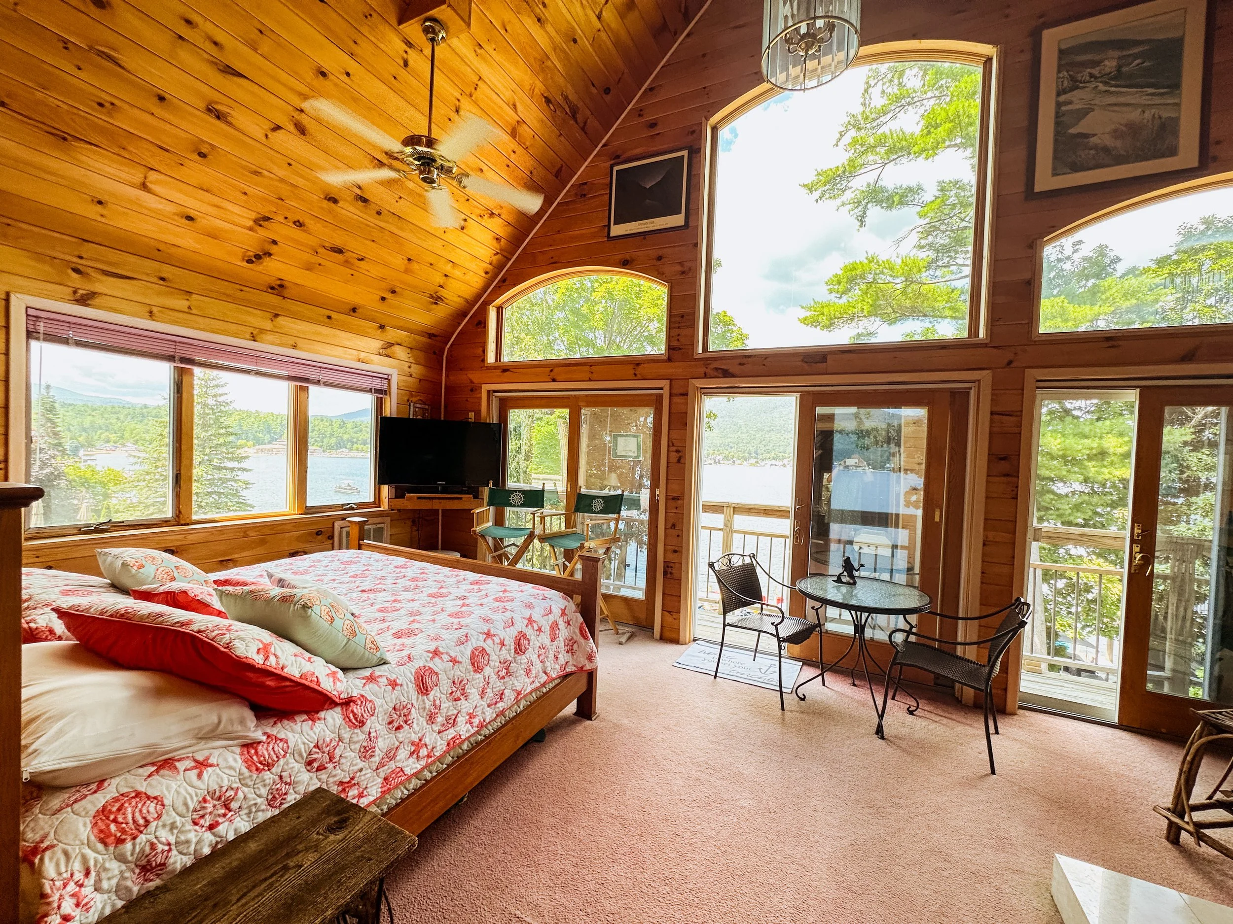 Living room with wood-paneled vaulted ceiling, large windows showing a lake and trees, a queen-sized bed with seashell-patterned quilt, and small sitting area with table and chairs.
