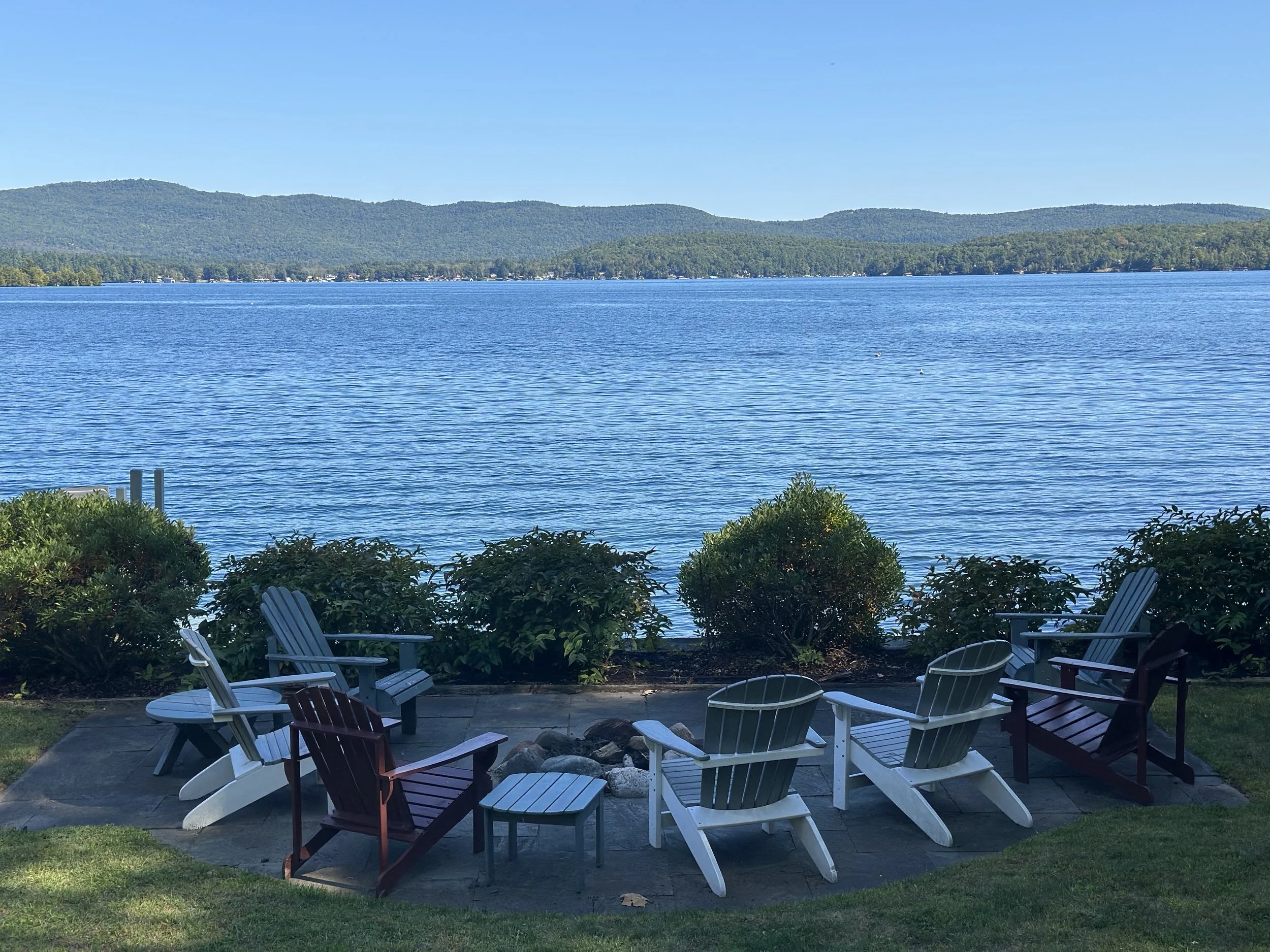 A lakeside view with Adirondack chairs arranged around a small table on a stone patio, overlooking a calm lake with distant mountains under a clear blue sky.