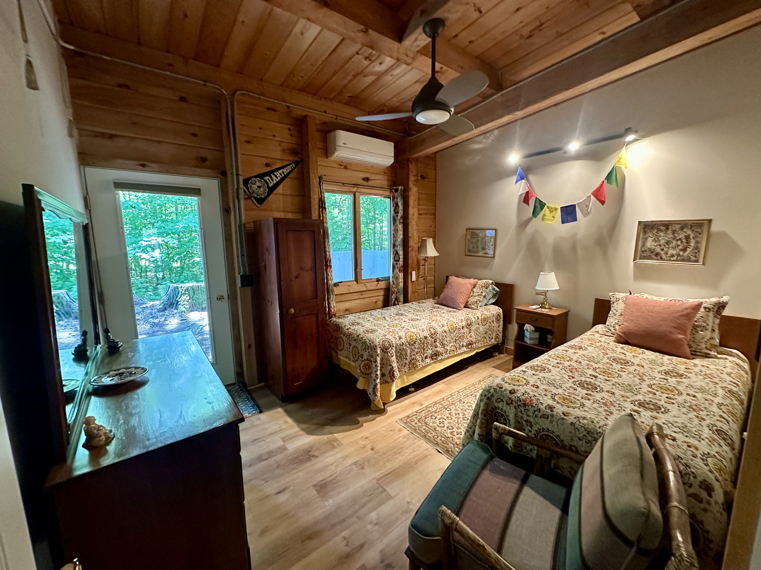 A cozy bedroom with wooden walls and ceiling, two single beds with floral bedspreads and pillows, a small wooden nightstand, a lamp, a colorful banner, and a window showing a wooded outdoor scene.