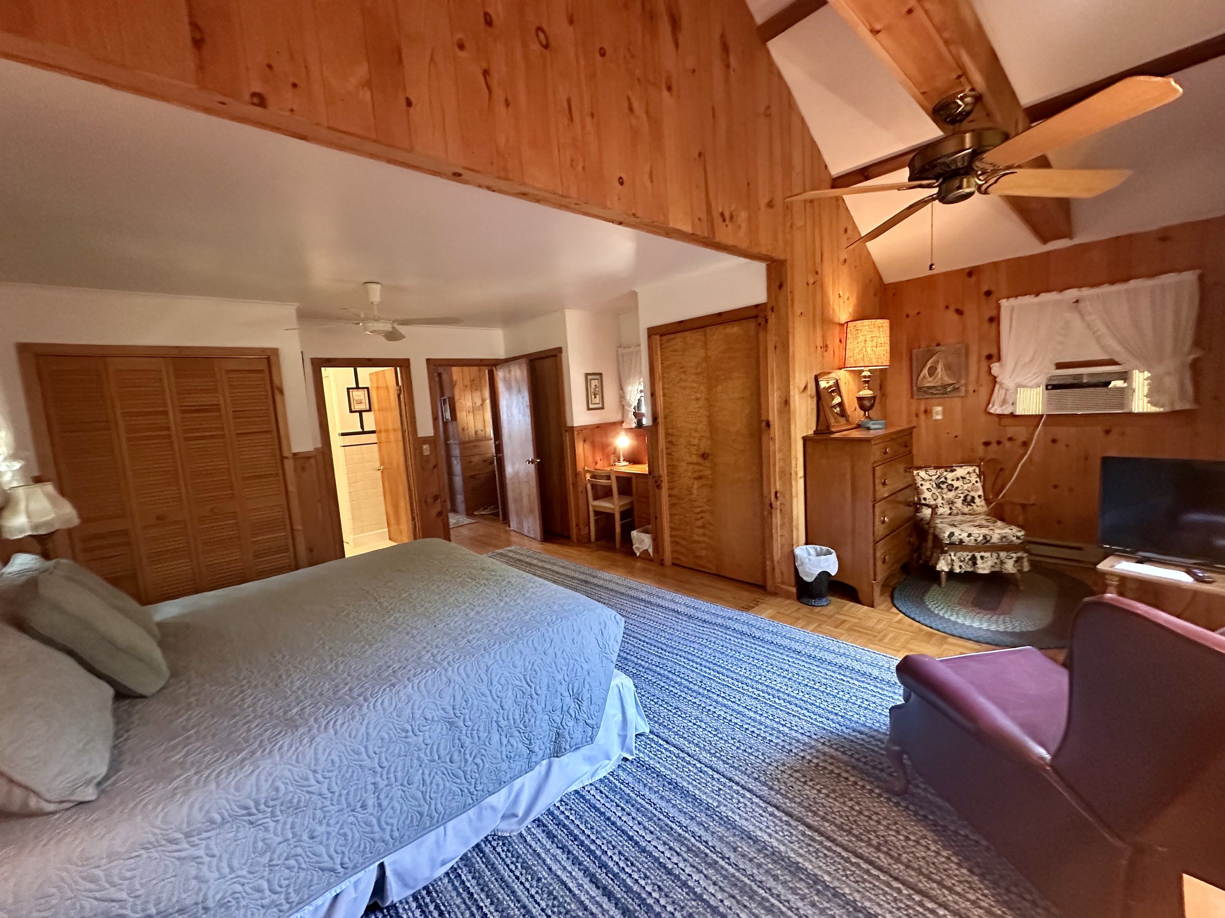 A cozy bedroom with wooden walls, a large bed with pillows, a ceiling fan, a dresser with a lamp, an armchair, a small TV, and a window with curtains.