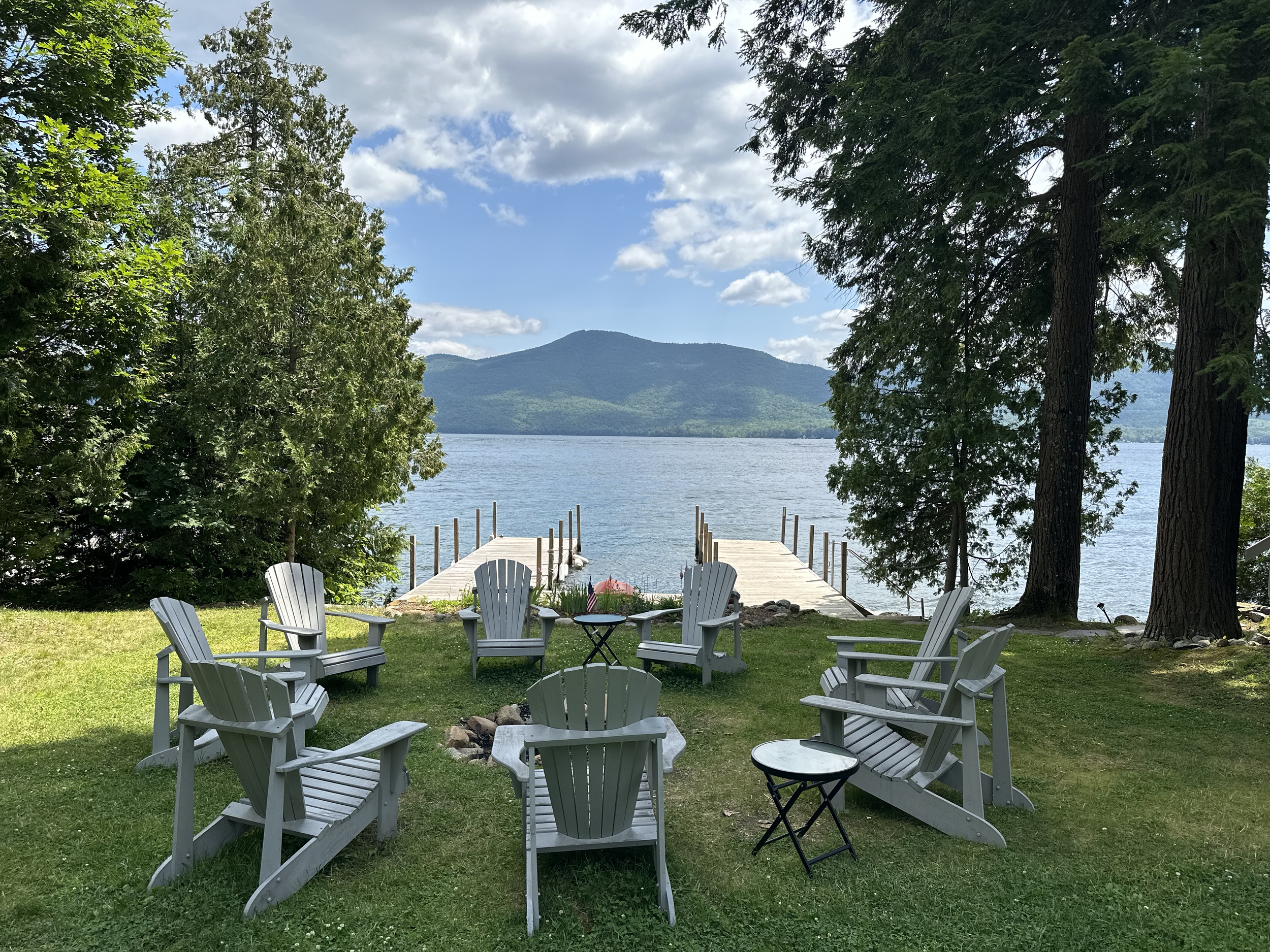 Seating area with eight white Adirondack chairs arranged in a circle outdoors on a grassy lawn overlooking a body of water with two docks, tall trees, and mountains in the background.