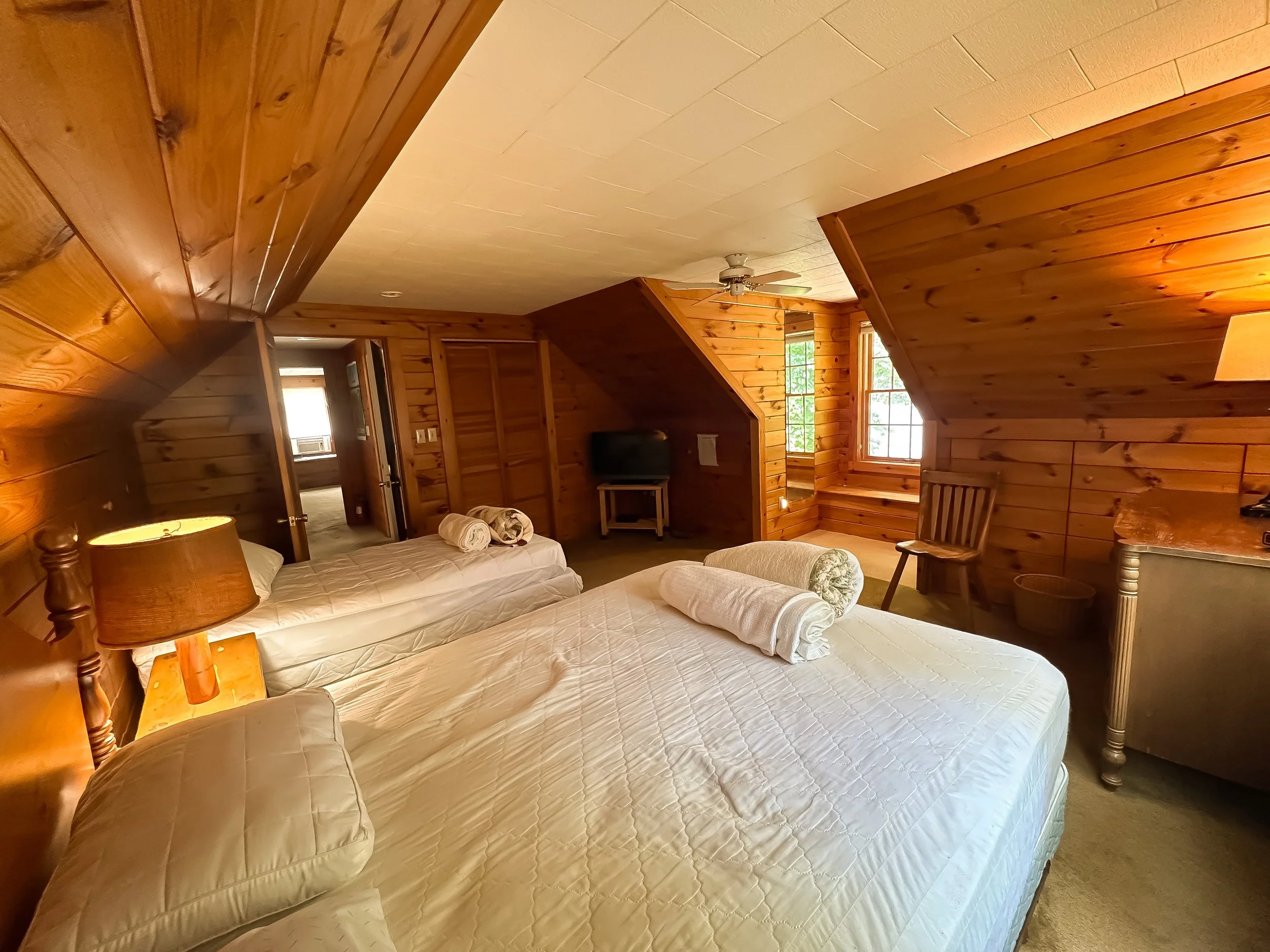 A rustic wooden bedroom with two beds, each with rolled towels, a small TV, a wooden chair, and windows letting in natural light.