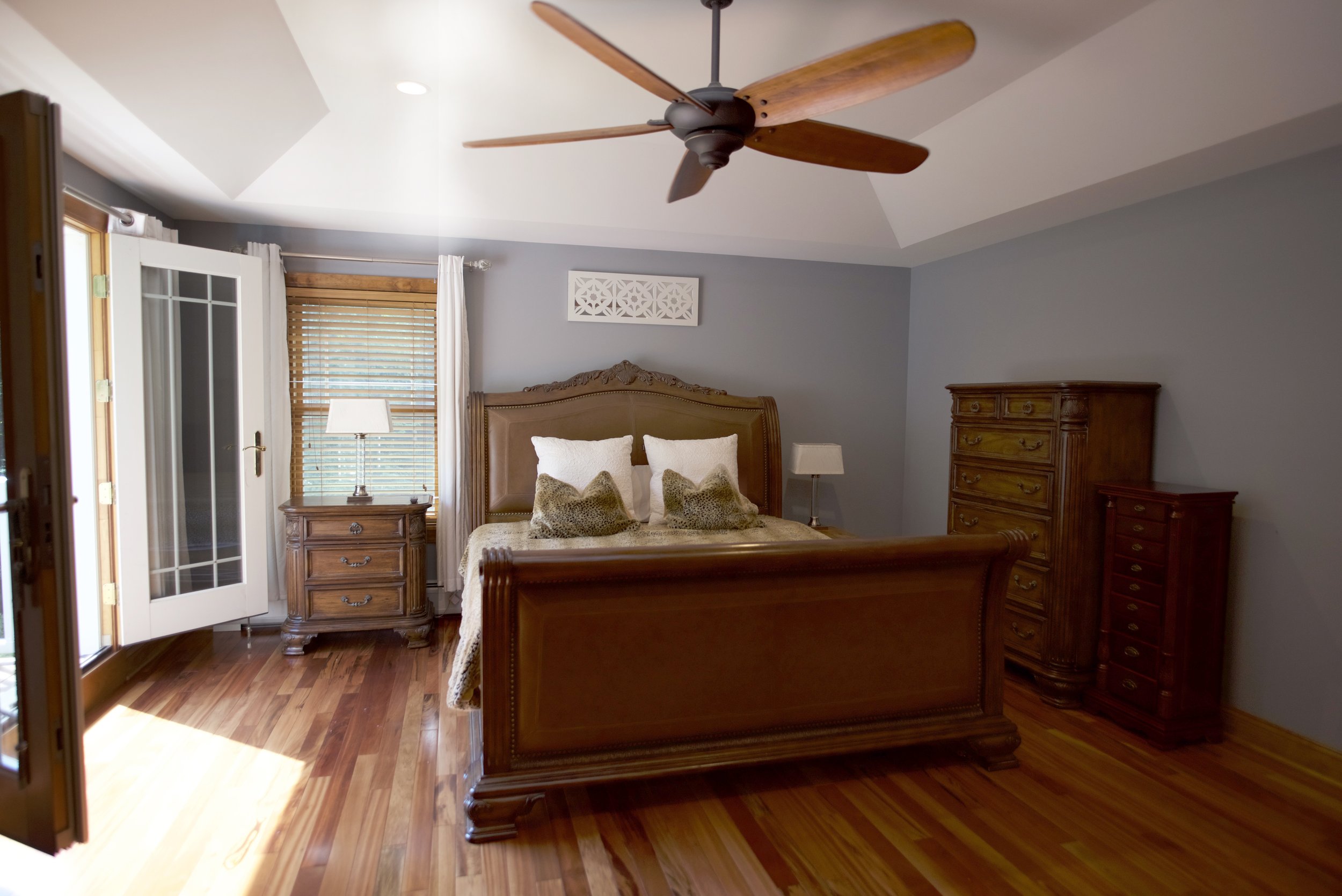 Bedroom with wooden bed frame, dresser, nightstand, ceiling fan, hardwood floors, gray walls, white curtains, and open door with blinds.