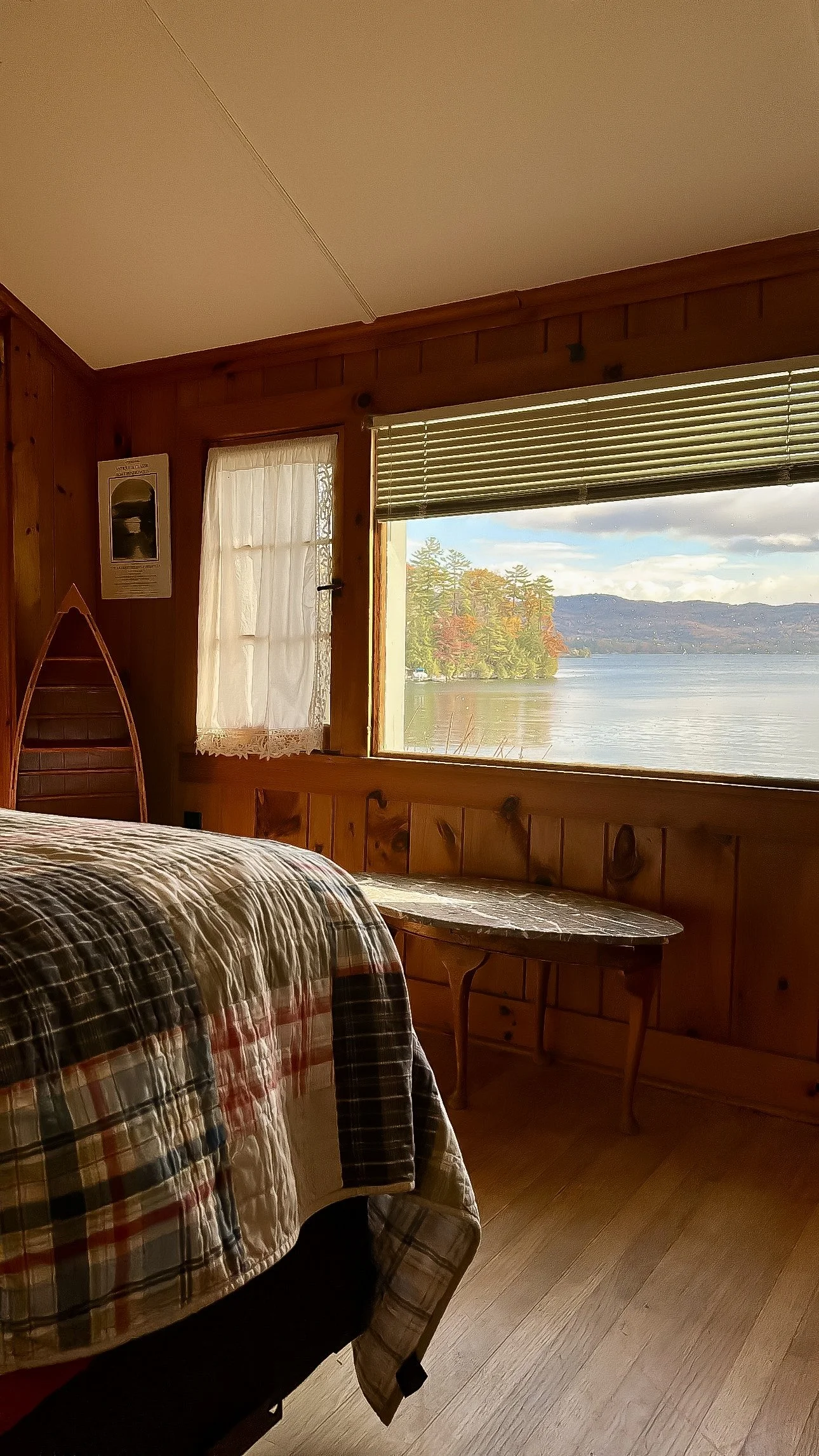A cozy wooden cabin bedroom with a bed covered in a plaid quilt, a window with partial blinds showing a lakeside view with trees and hills in the background, a small table beside the bed, and a curtain on a smaller window.