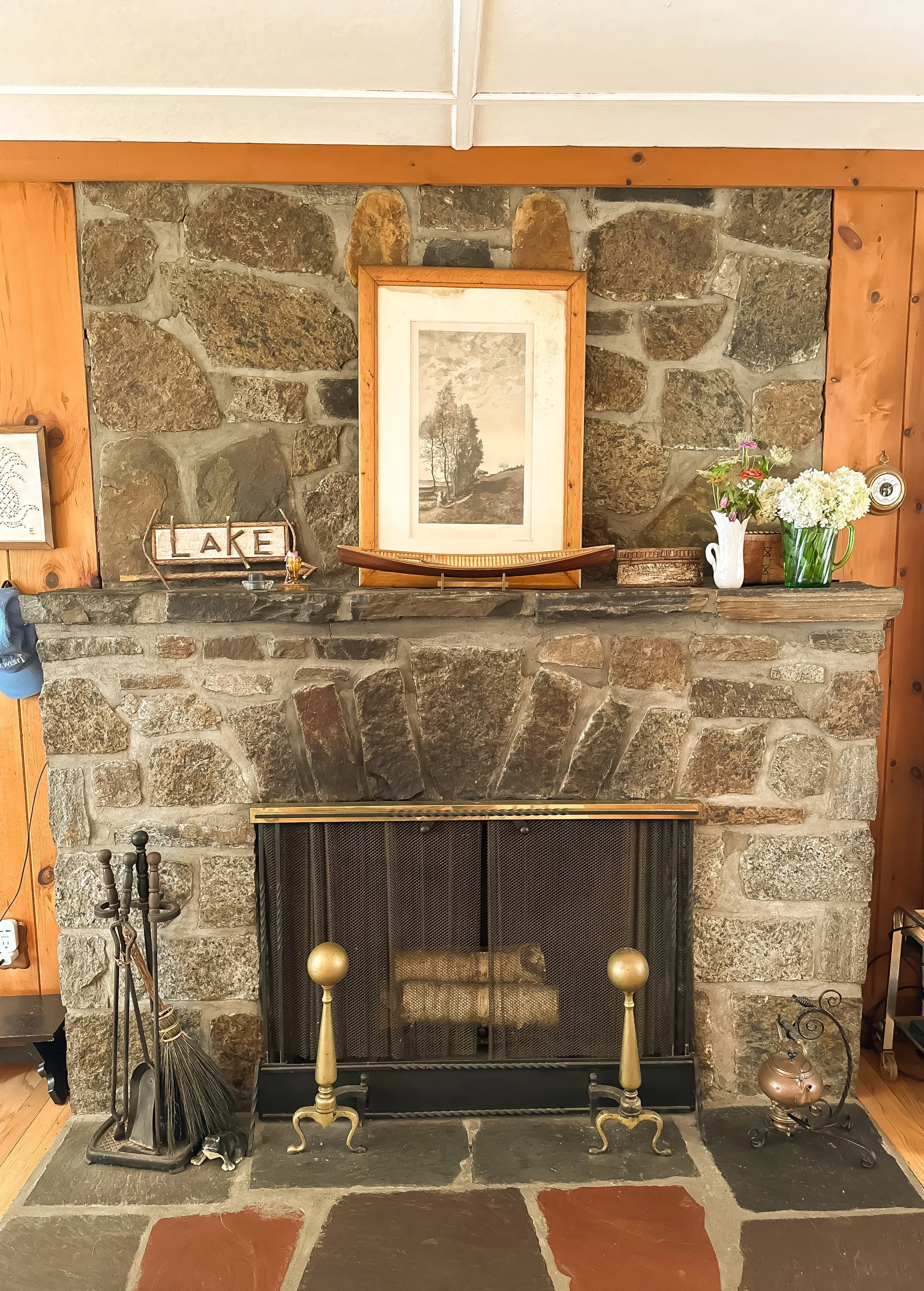 A rustic stone fireplace with a wooden mantel decorated with a framed black-and-white landscape drawing, a small boat model, a green vase with white flowers, and a white jug with pink and white flowers. The fireplace has brass and iron tools on eithe