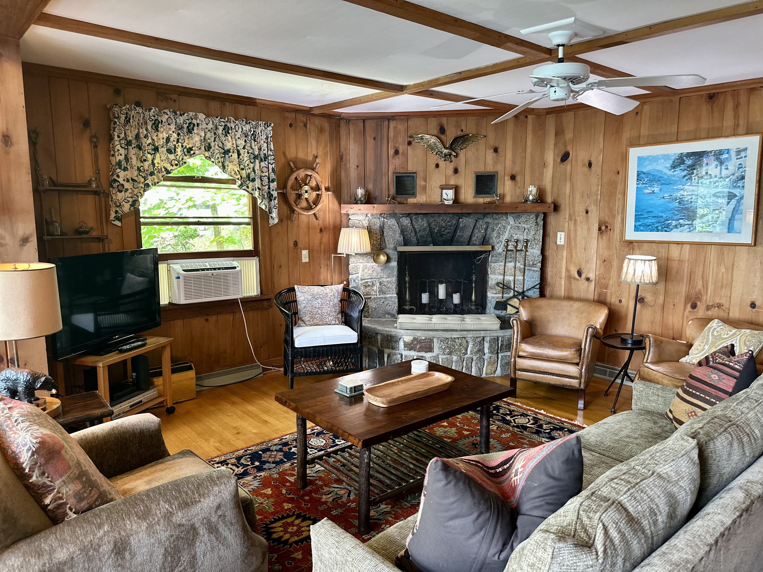 Cozy living room with wood-paneled walls, a stone fireplace, a ceiling fan, a wooden coffee table, a sofa, armchairs, a television, and decorative items.