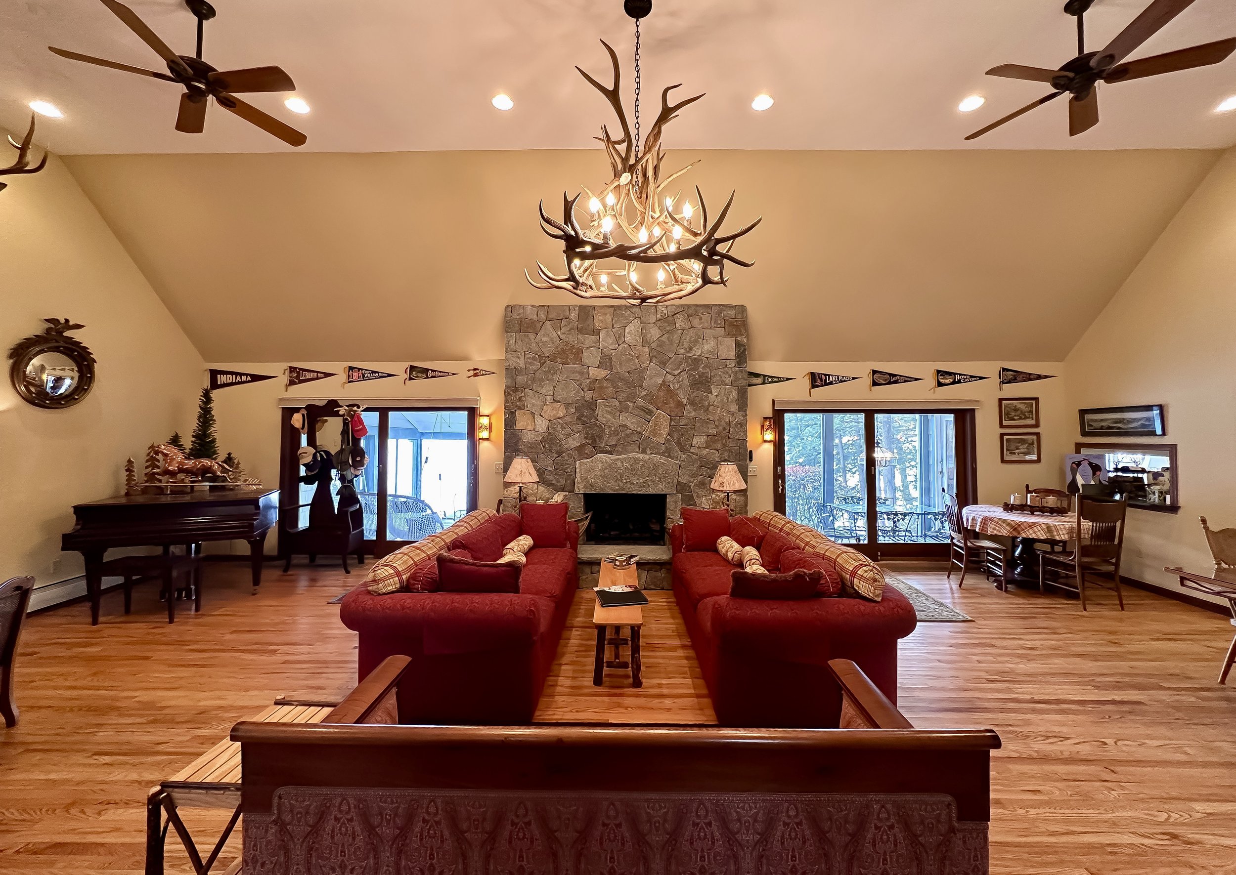 A spacious living room with a high vaulted ceiling, two red sofas facing a stone fireplace, and an antler chandelier hanging overhead. There are framed pennants on the walls, a grand piano on the left, and a dining area on the right, with sliding gla