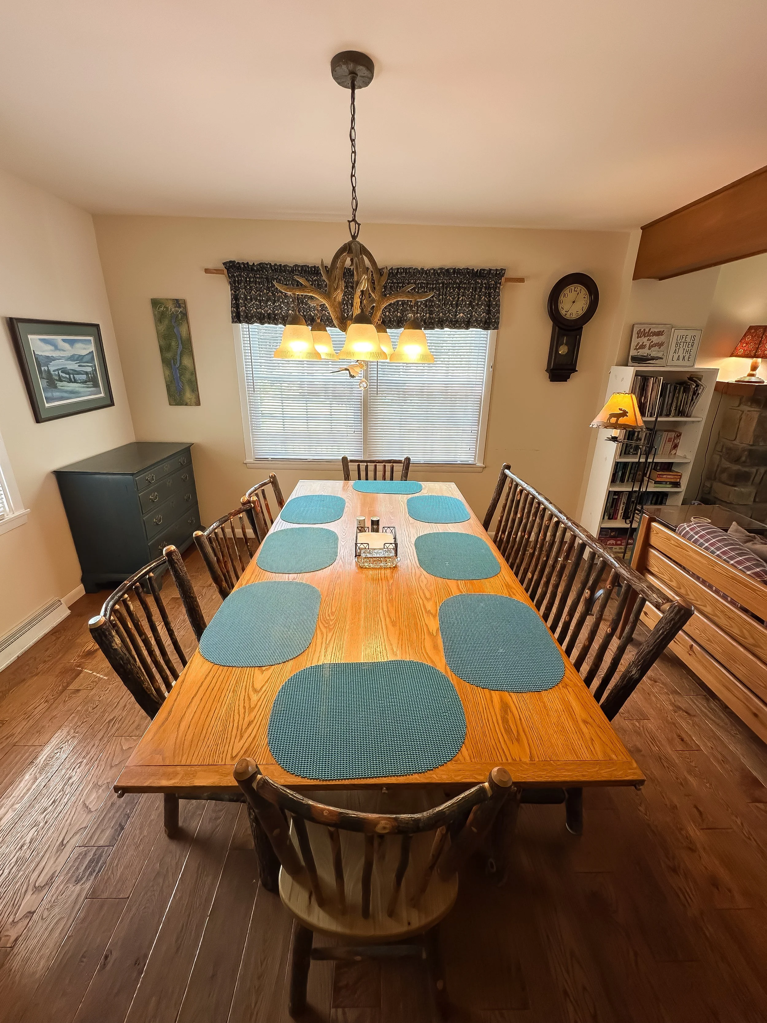 Dining room with a wooden table set with ten teal placemats, surrounded by wooden chairs, illuminated by a chandelier with a branch design, and a large window with blinds and a floral valance. Additional furniture includes a chest of drawers and a bo