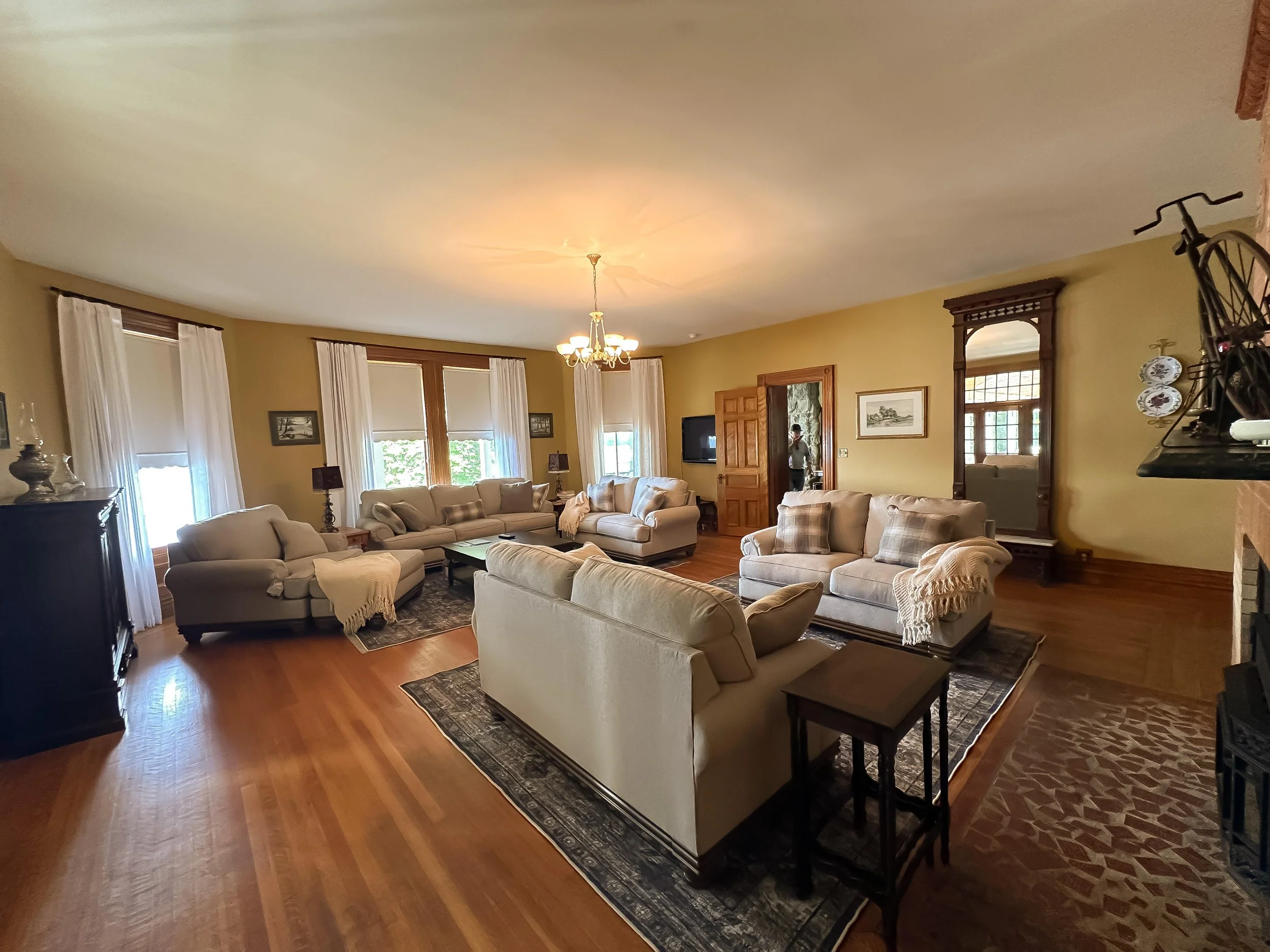Living room with beige sofas, wooden floor, large windows with white curtains, chandelier, and a person standing near a doorway.