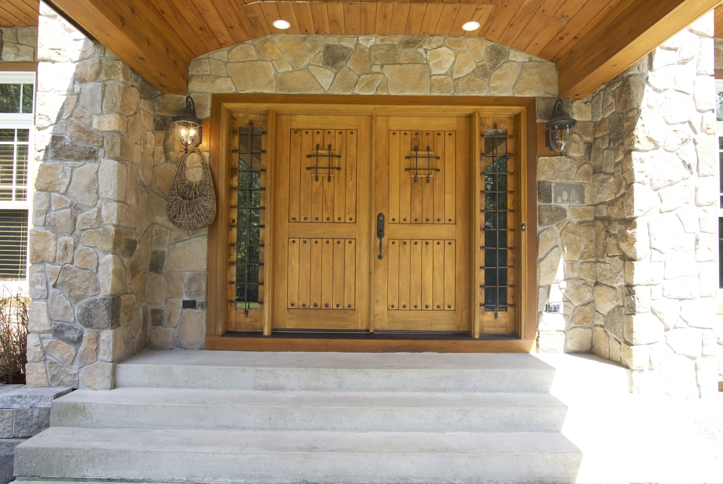 Wooden front door with black hardware, flanked by stone walls and glass sidelights, under a wooden ceiling, with two outdoor wall lanterns on either side.