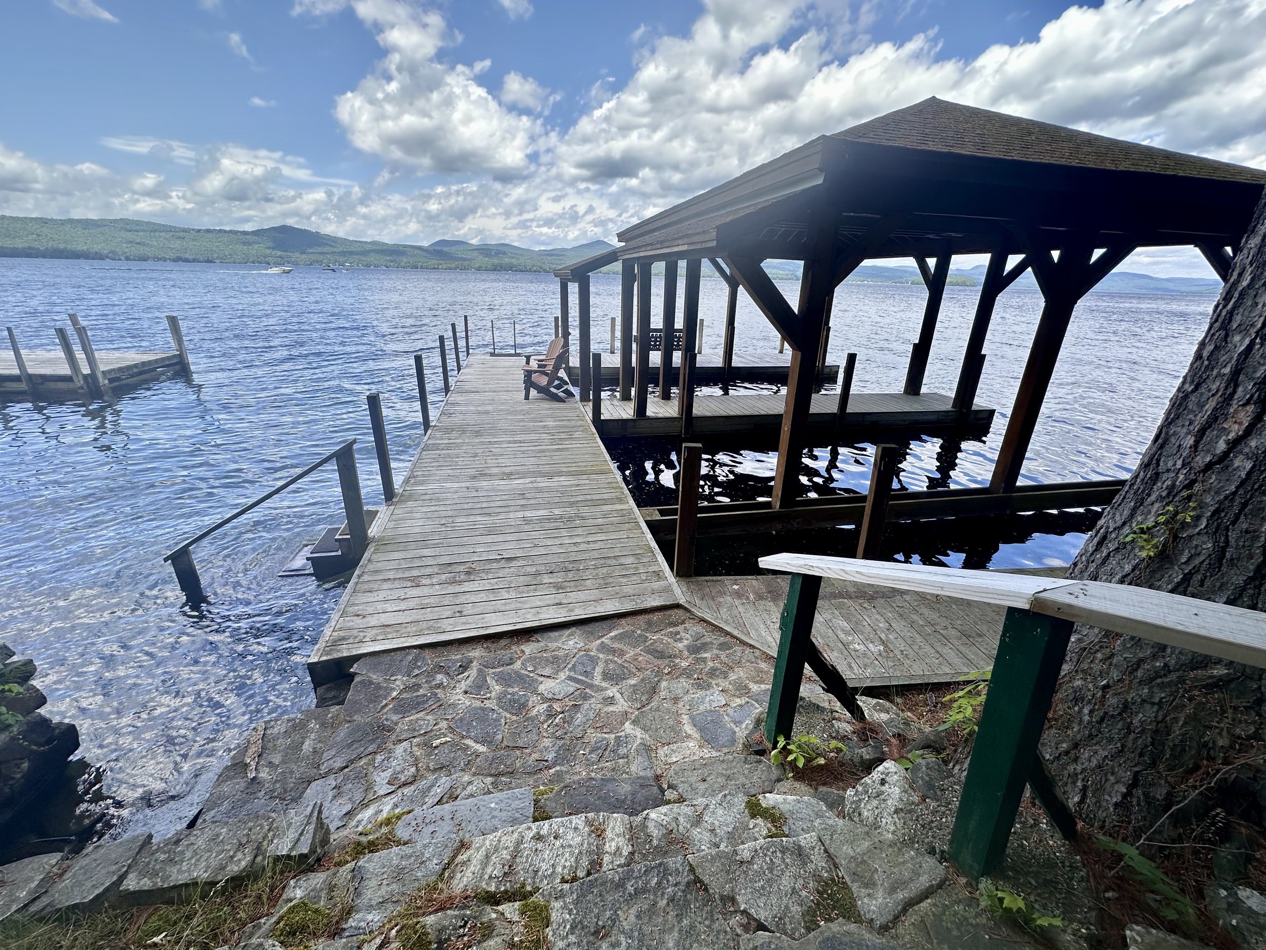 Wooden dock and gazebo on a lake with rocky shoreline, surrounded by mountains, under partly cloudy sky.