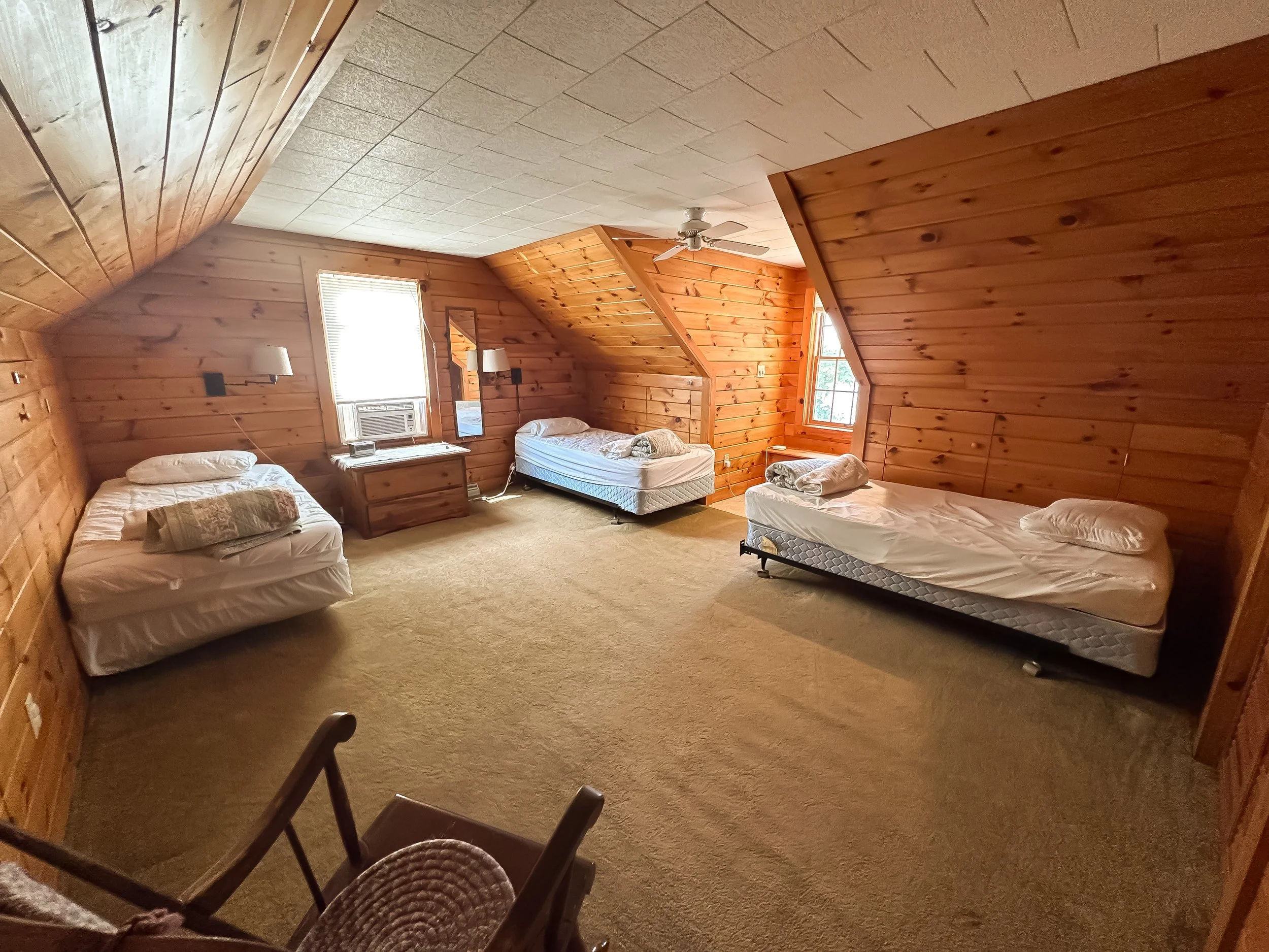 A spacious attic bedroom with wood-paneled walls and ceiling, three beds with white sheets and pillows, a small wooden dresser, three windows letting in natural light, a ceiling fan, and a carpeted floor.