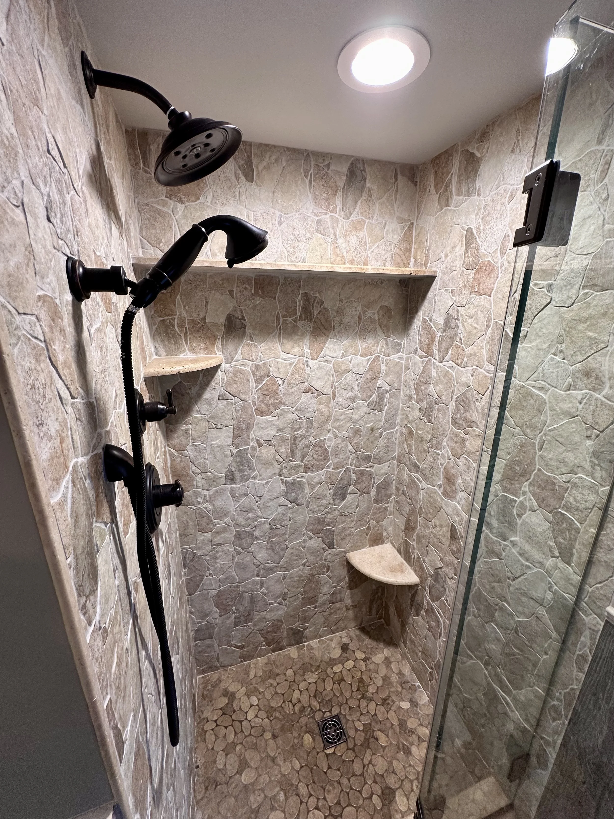 A shower with stone tiles, a built-in corner seat, and two black showerheads, one handheld and one fixed, with a glass door and ceiling light.