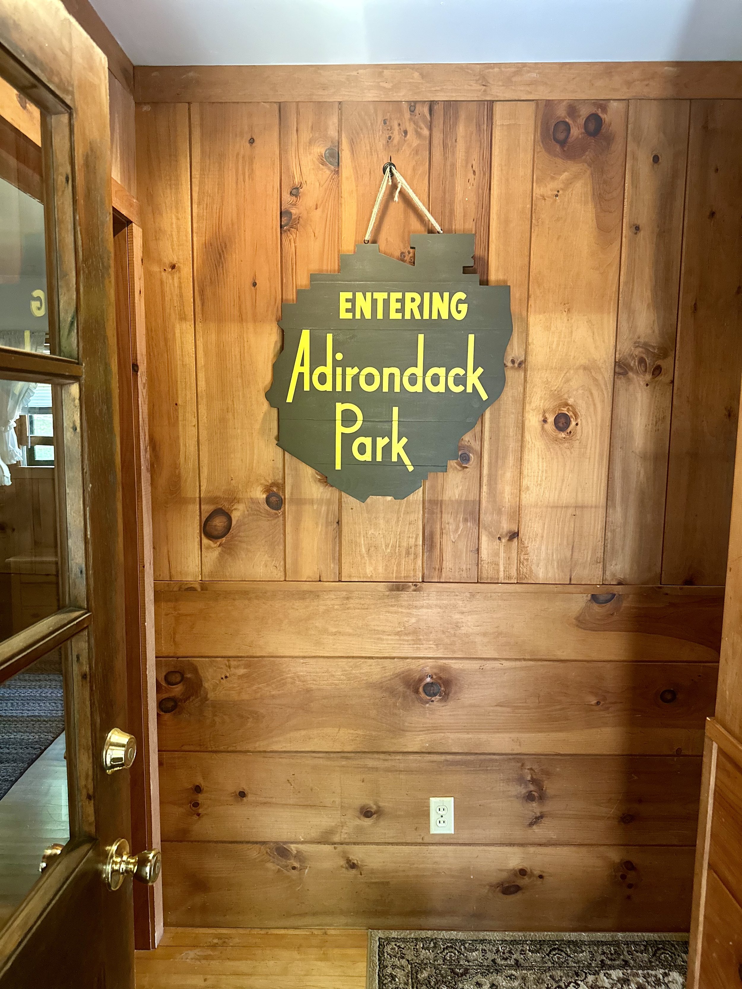 Wood-paneled wall with a sign that reads 'Entering Adirondack Park' in yellow lettering on a dark background.