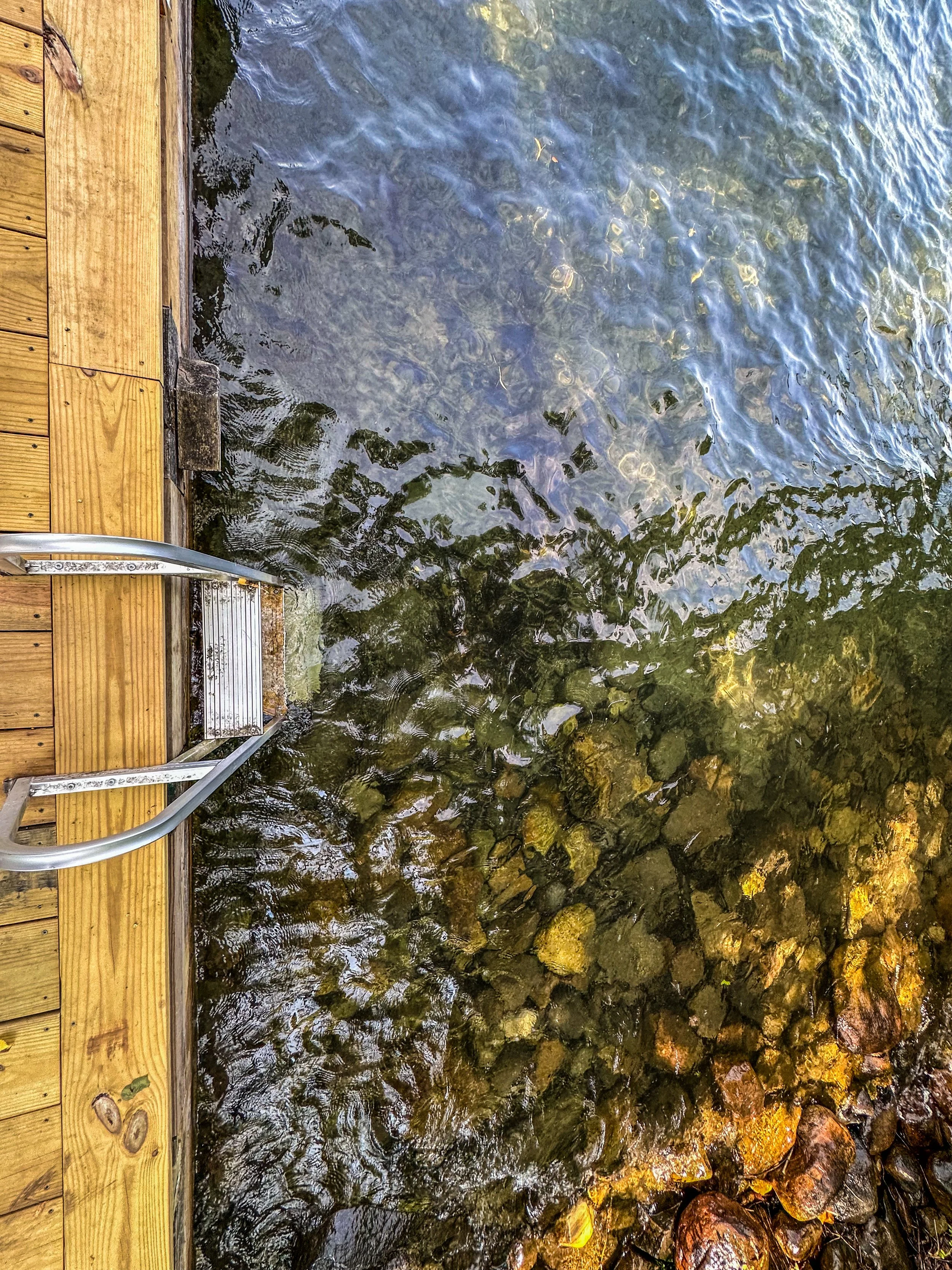 Wooden dock with metal ladder leading into clear water over rocks.