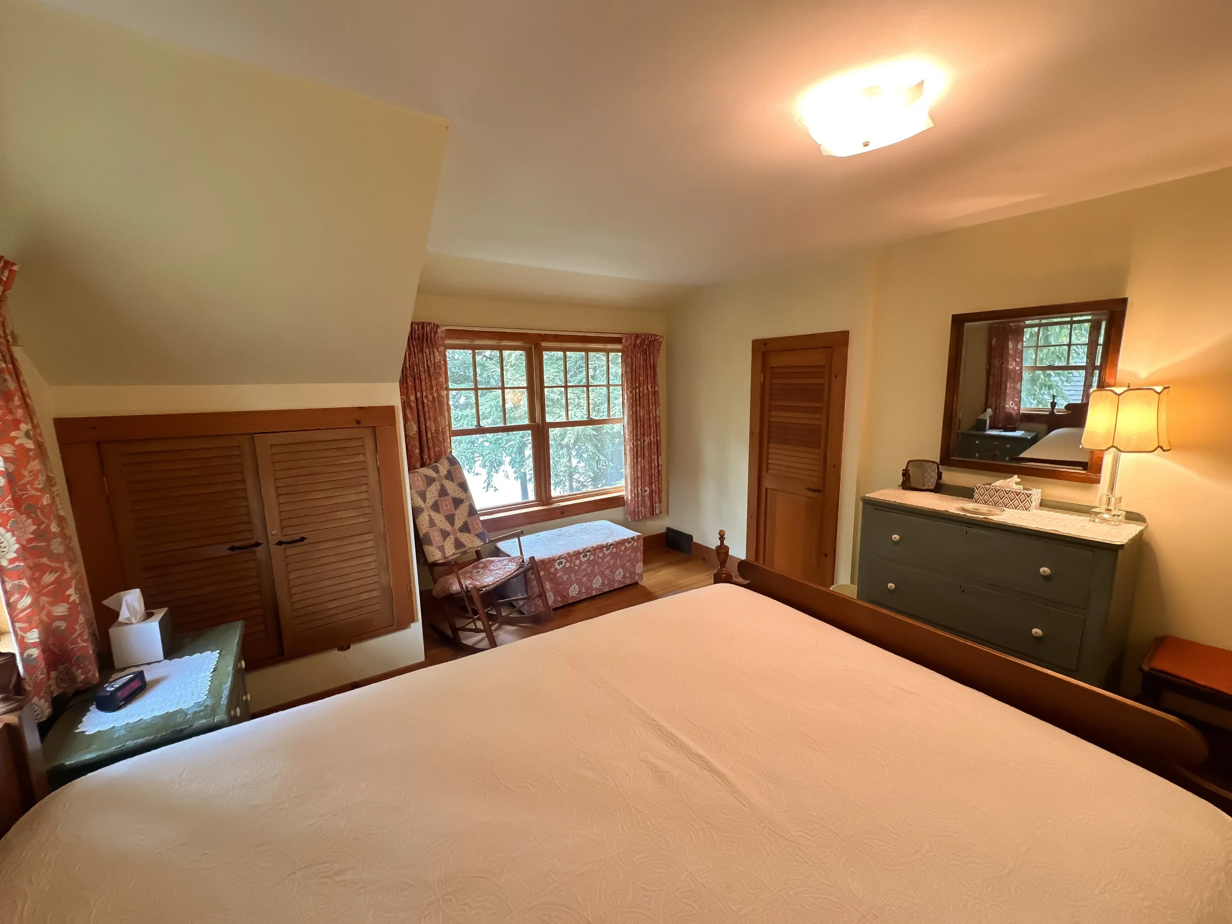 Cozy bedroom with a large bed, wooden furniture, floral curtains, and positioned near a window with a view of trees.