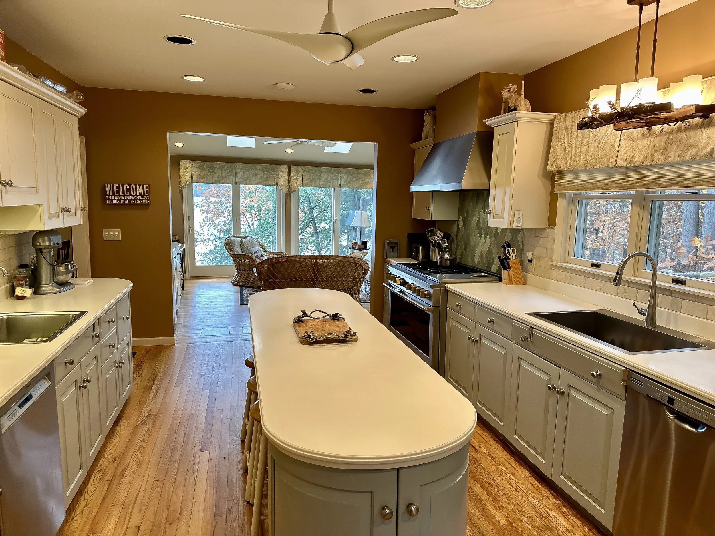 View of a kitchen with white cabinets, a large island with a tray, a beige wall, and a window above the sink. In the background, there is a living room area with large windows and a sitting area with wicker furniture.