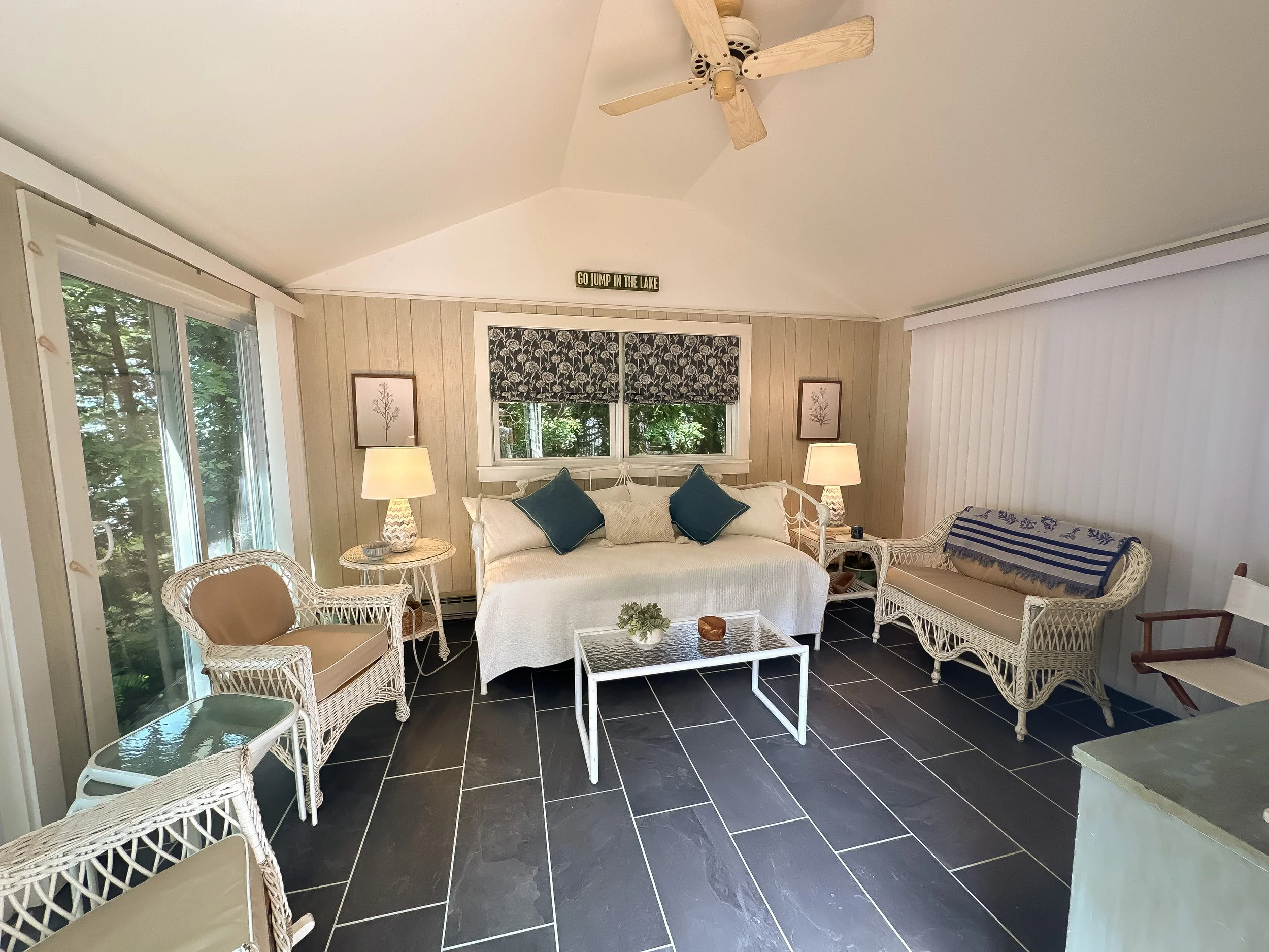 Living room with a white sofa, wicker chairs, and a black tiled floor, decorated with lamps, framed pictures, and a window with patterned curtains.