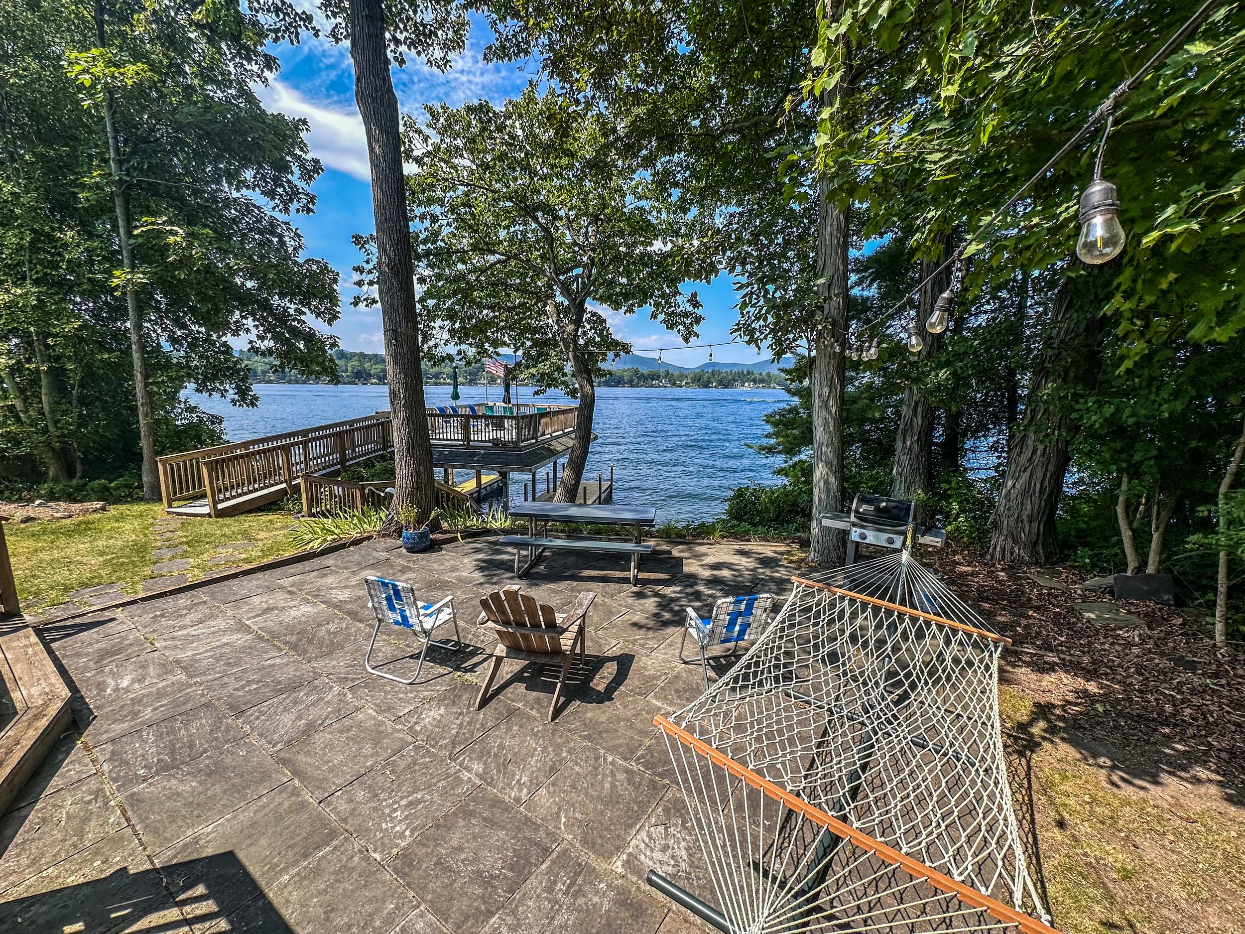 An outdoor patio area near a lake with a hanging hammock, outdoor seating, a barbecue grill, string lights, trees, and a wooden dock extending into the water under a partly cloudy sky.