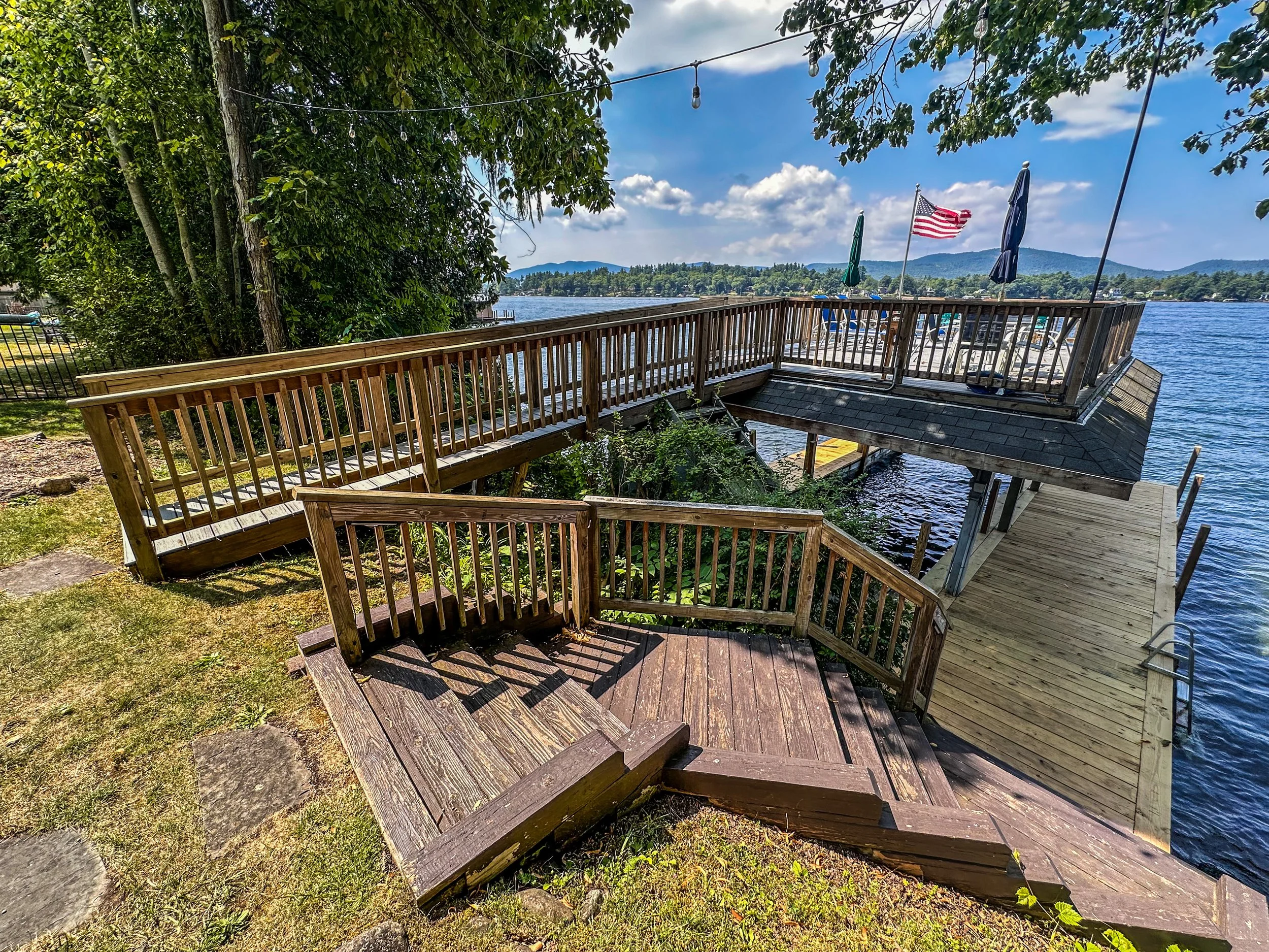 View of a wooden deck and stairs leading to a boat dock that extends over a body of water. There are umbrellas and an American flag on the deck, with trees on one side and hills in the background under a partly cloudy sky.