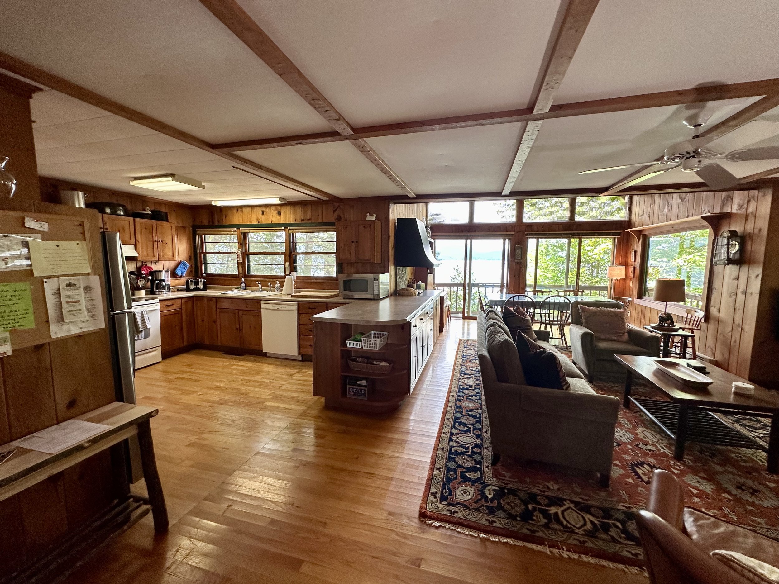 Open-concept living room and kitchen area with wood-paneled walls and ceiling, hardwood floors, large windows providing natural light, a sofa and armchair arrangement, and a view of trees and water outside.