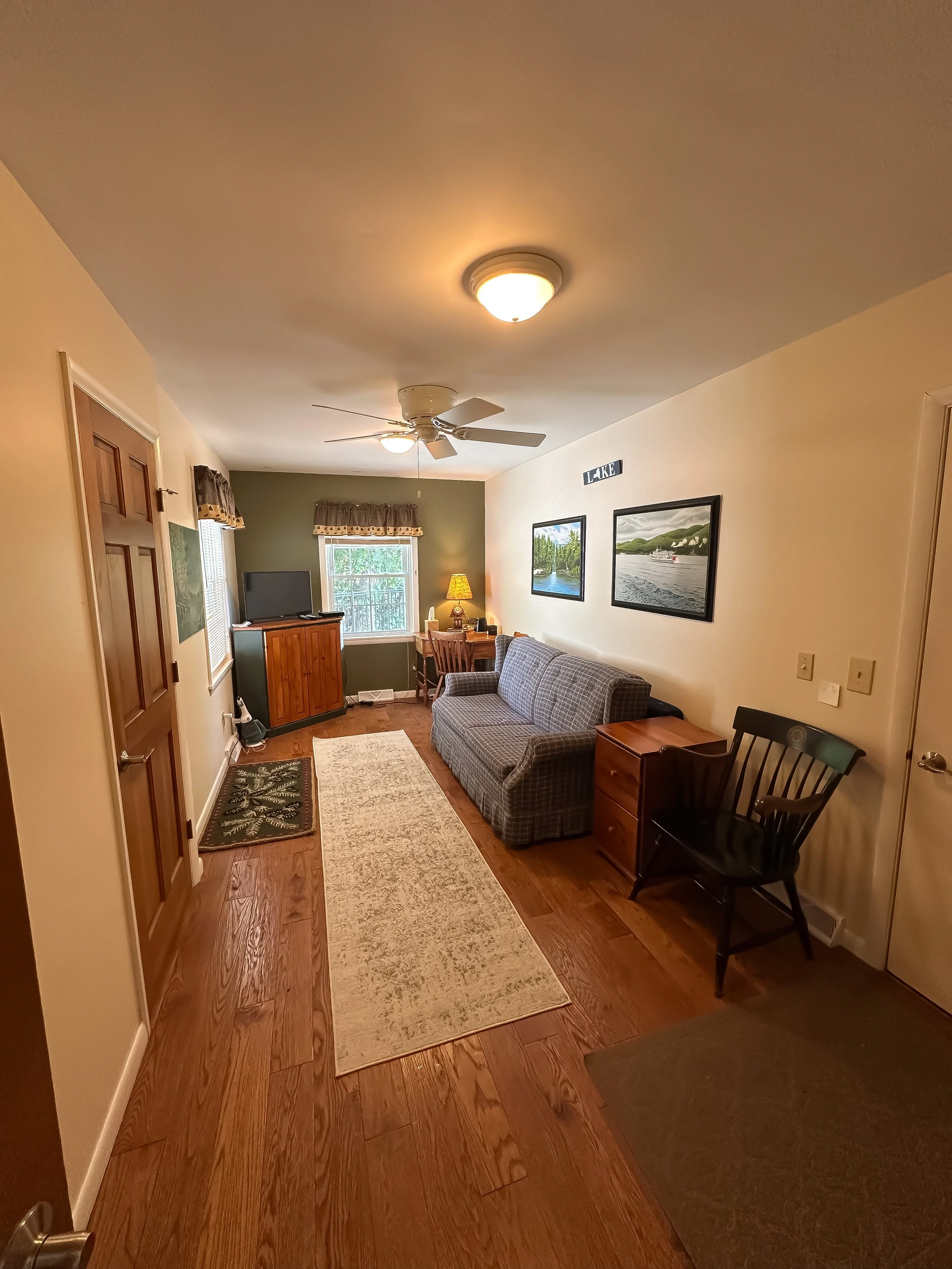 Living room with wooden floor, beige walls, a plaid sofa, a black chair, a small wooden side table, and a TV stand with a TV. There are picture frames on the wall, a window with patterned curtains, a ceiling fan, and a rug.