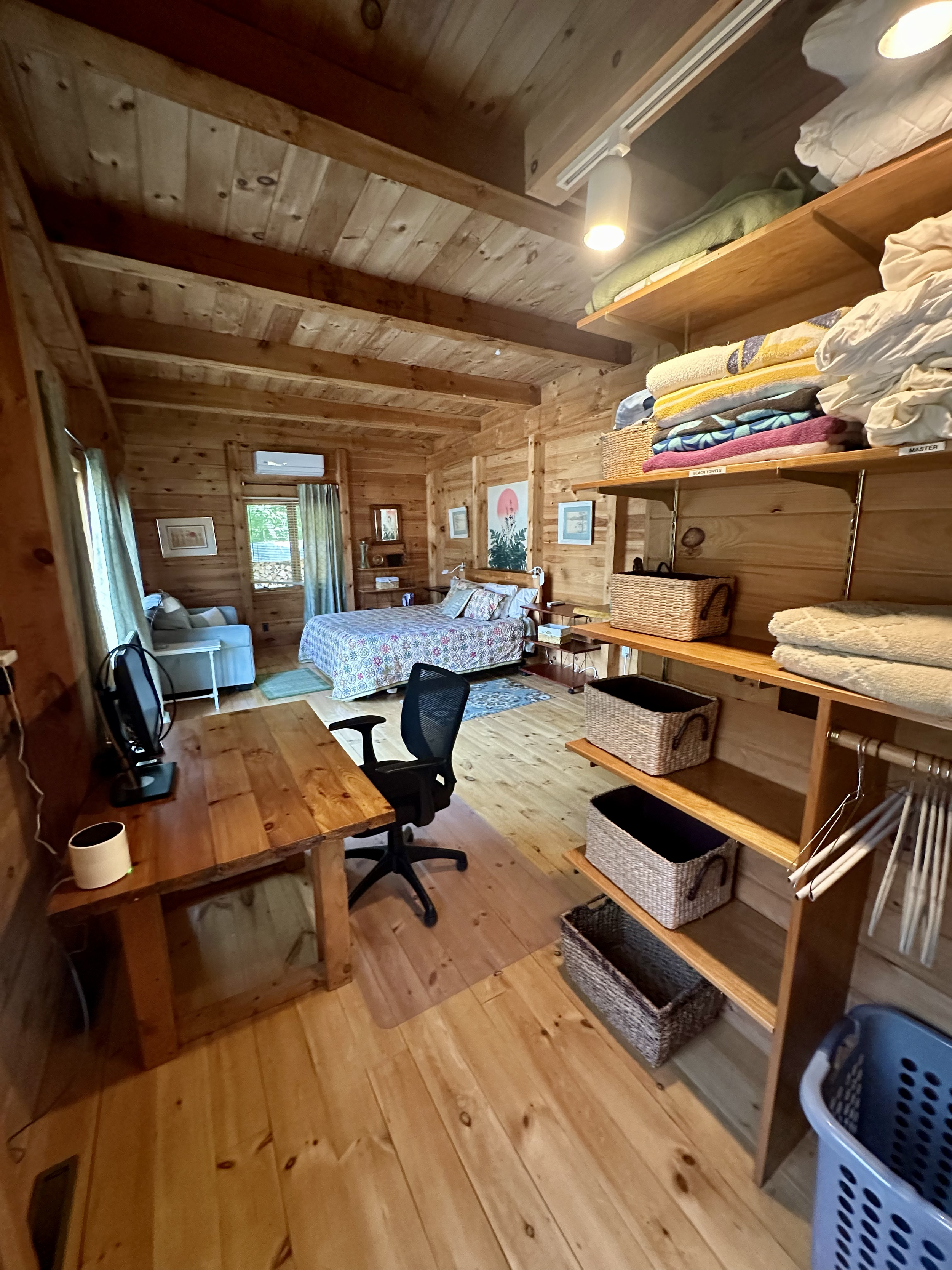 A cozy wooden bedroom with a bed, workspace, and organized shelves with folded towels and baskets.