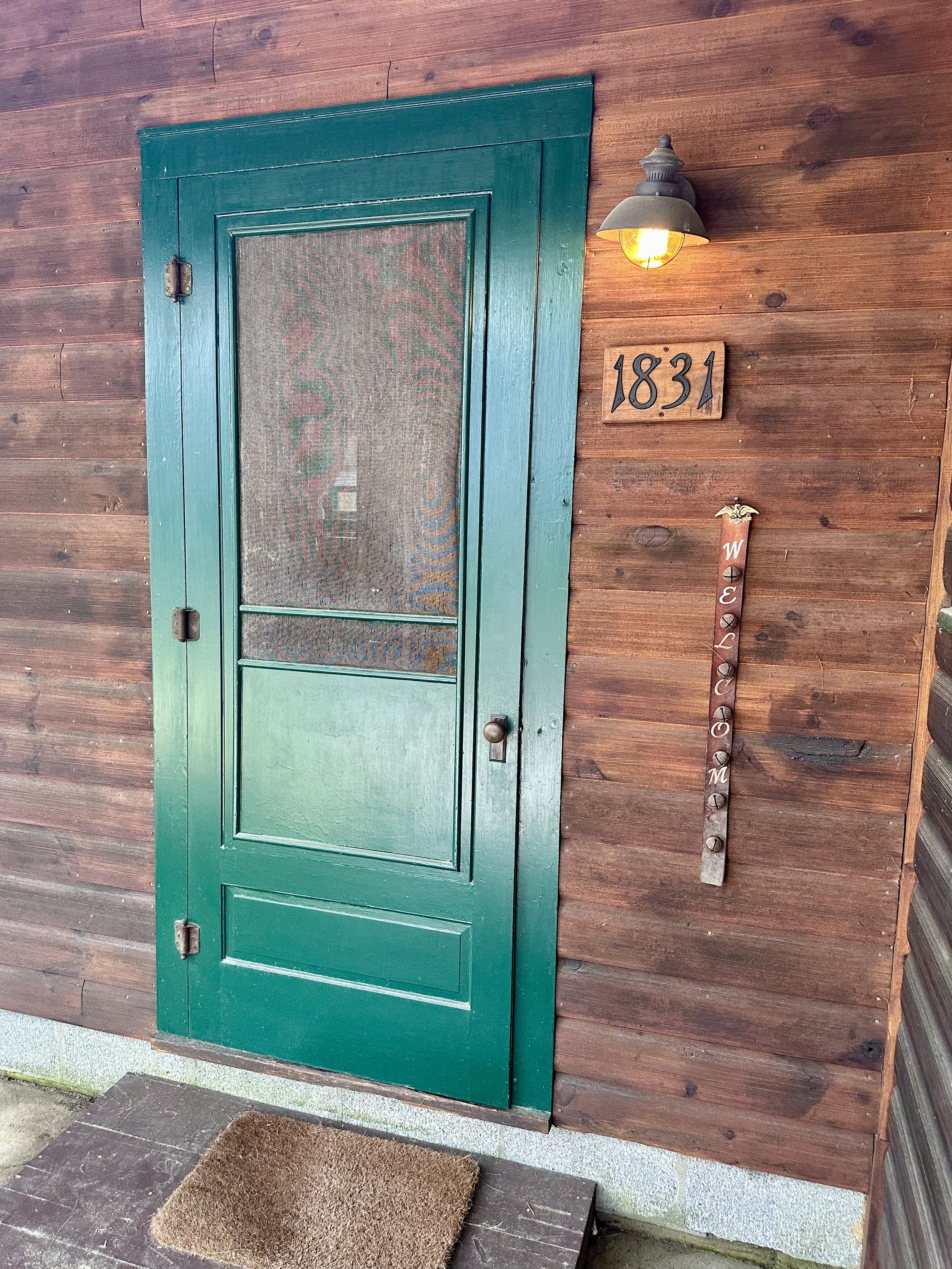 Front door of a house with a green wooden screen door, a wall-mounted light fixture, house number 1831 on the wall, and a decorative vertical sign with the word 'WELCOME' on it.