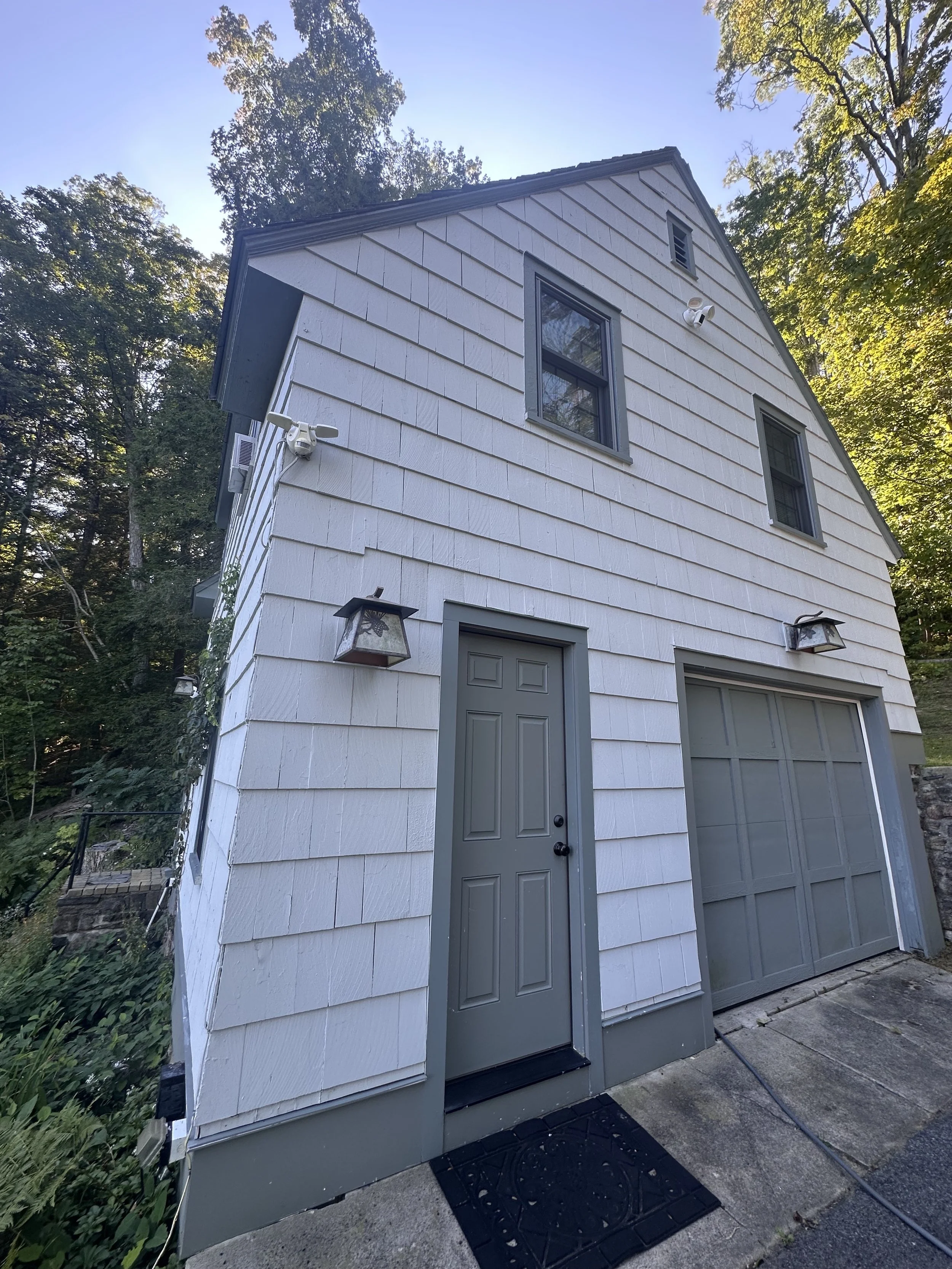 White two-story house with gray trim, two windows on the upper level, a gray front door, and a garage door. Surrounding trees and sunlight above.