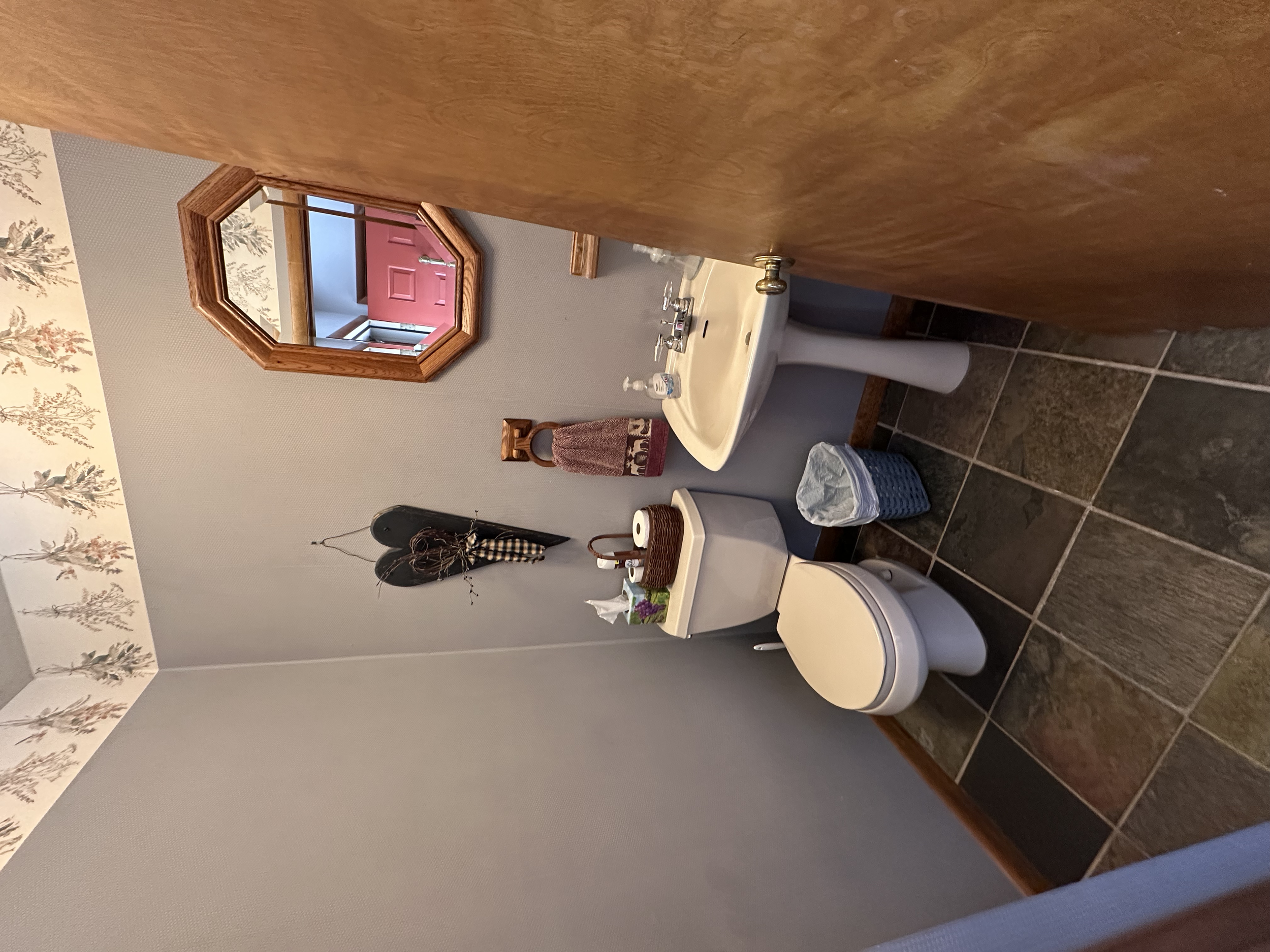 Bathroom with a toilet, pedestal sink, mirror, patterned wallpaper, and various decorations.