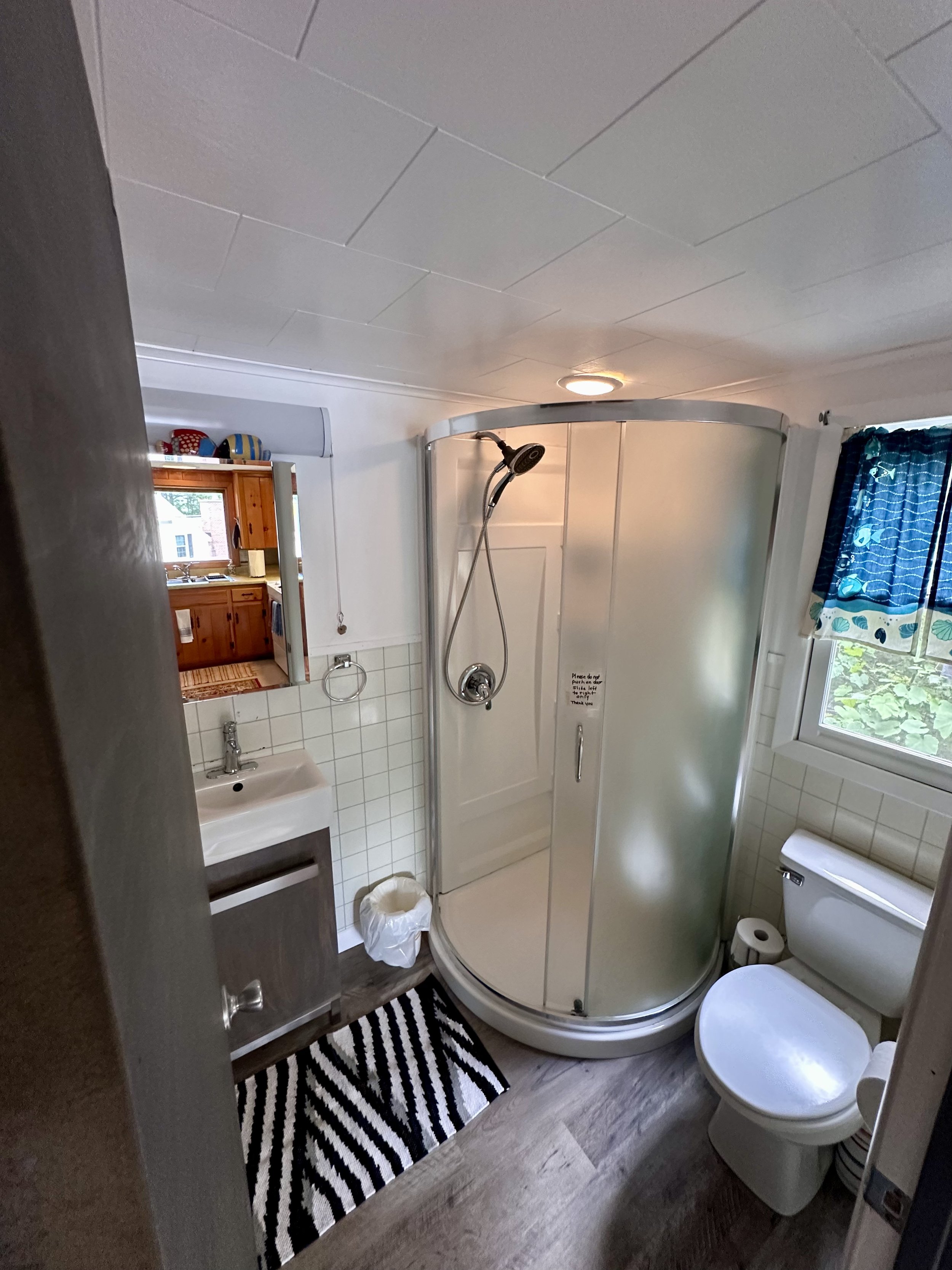 Small bathroom with a corner shower unit, toilet, small sink with cabinet, mirror, and window with blue curtain, wooden floor, and a black-and-white striped rug.