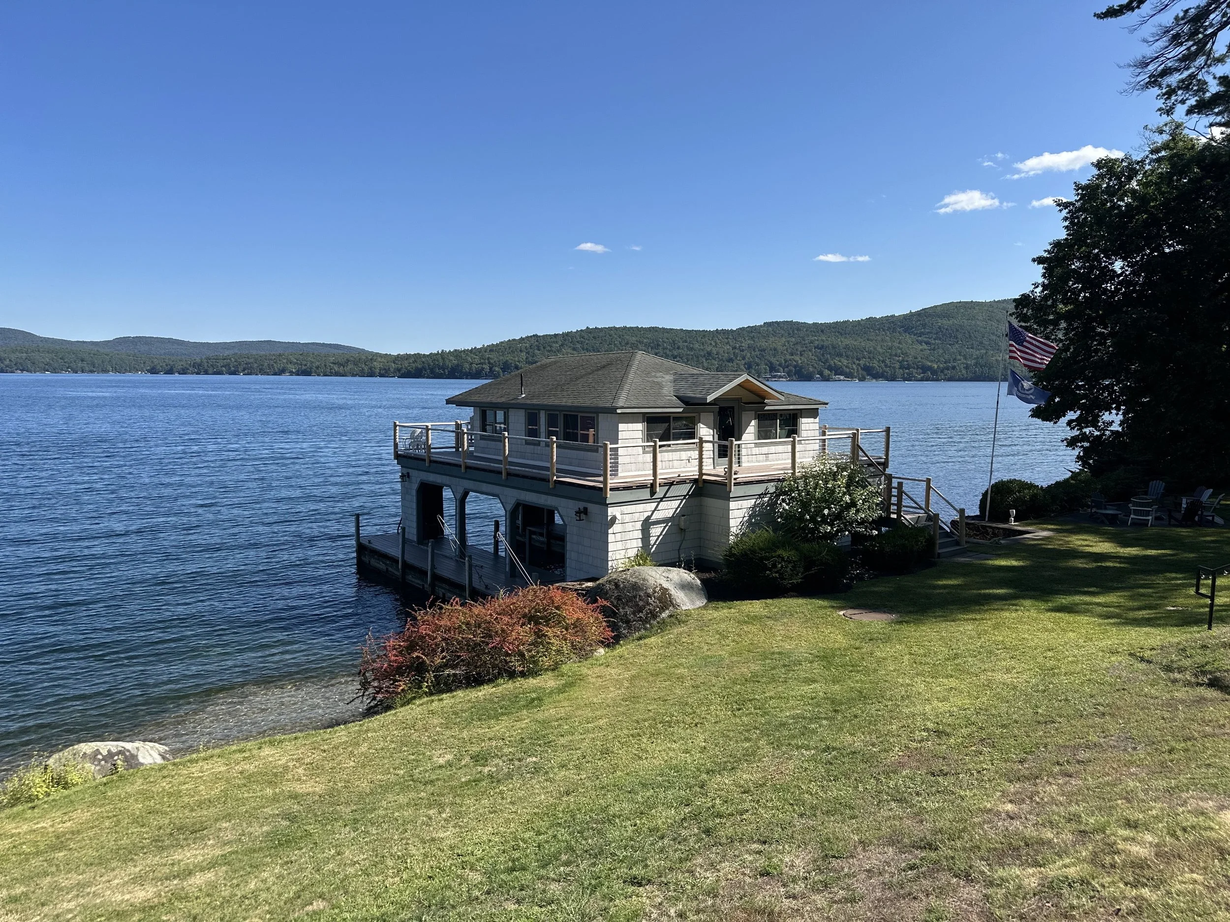 A house with a large deck built over the water on a lakeshore, surrounded by grass, bushes, and trees, with a flagpole and Adirondack chairs nearby.