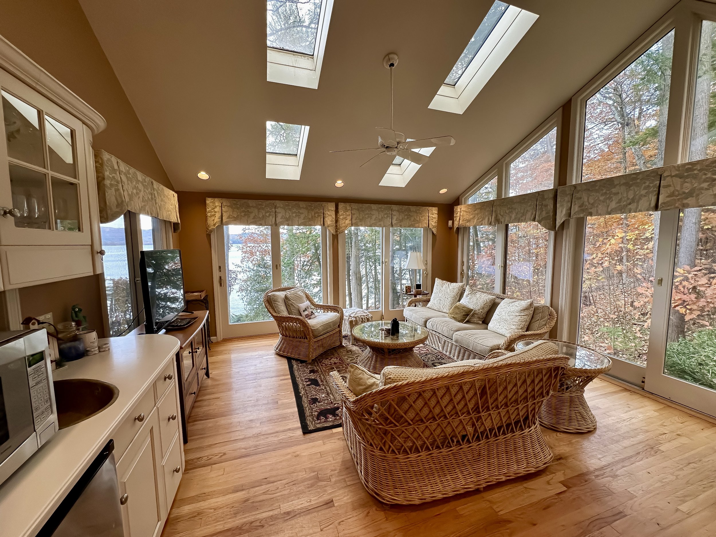 A sunroom with large windows and skylights, furnished with wicker chairs, a sofa, a glass coffee table, and a TV, surrounded by trees with autumn leaves outside.