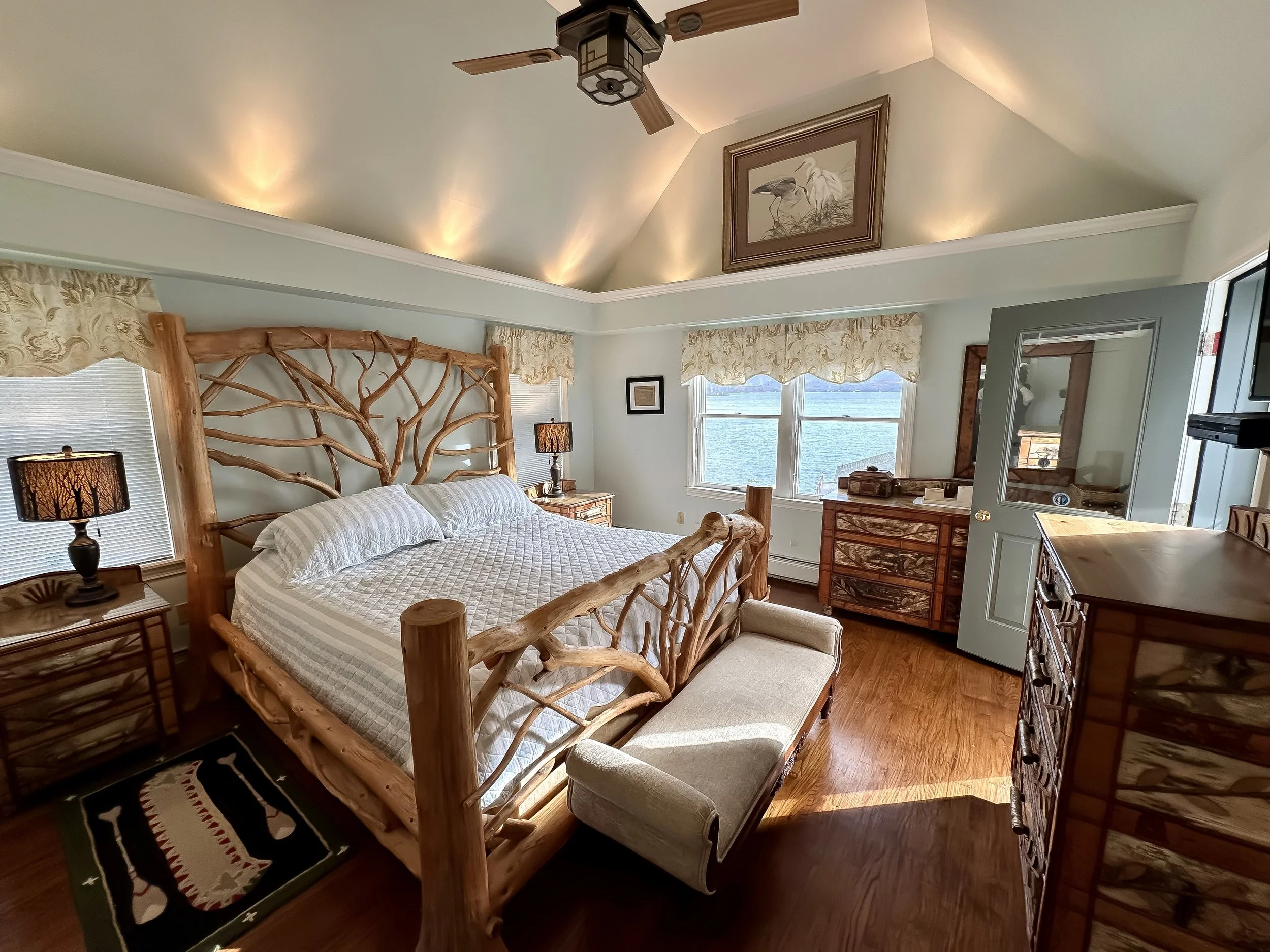 A bedroom featuring a large wooden bed with a branch-like headboard and footboard, two nightstands with lamps, a chest of drawers, a bench at the foot of the bed, a dresser with a mirror, three windows showing water view, and decorative artwork on th