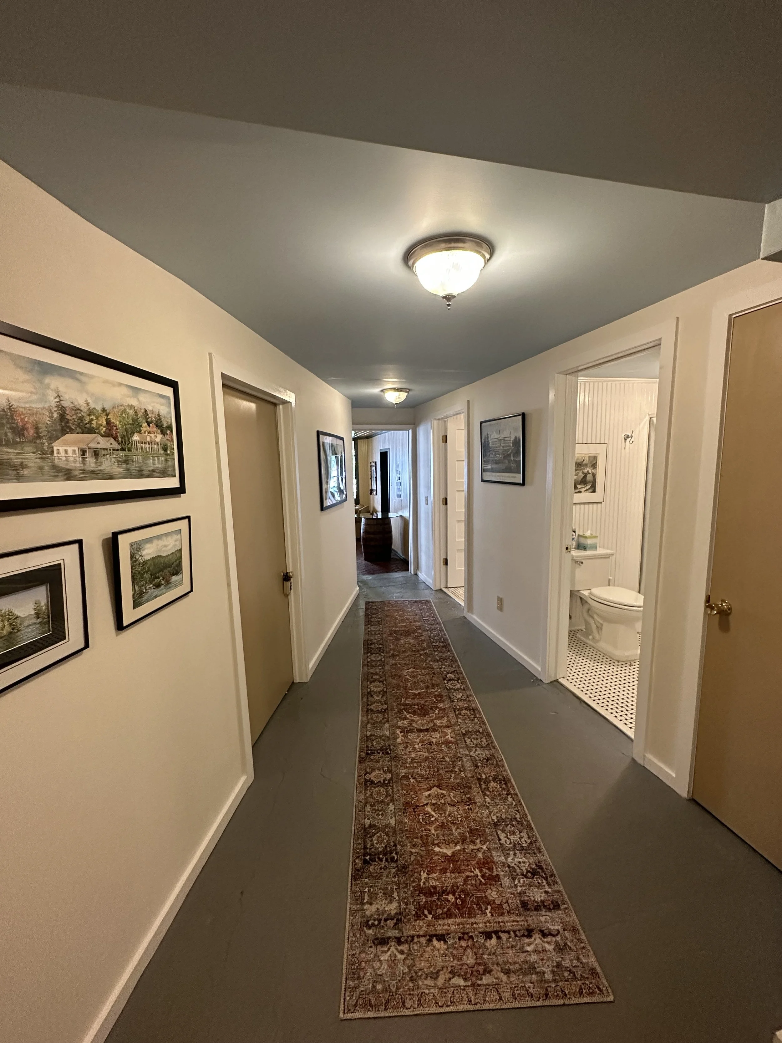 A hallway with cream walls and a long ornate rug, framed artwork on the walls, a doorway to a bathroom with black and white tile flooring, and ceiling lights.