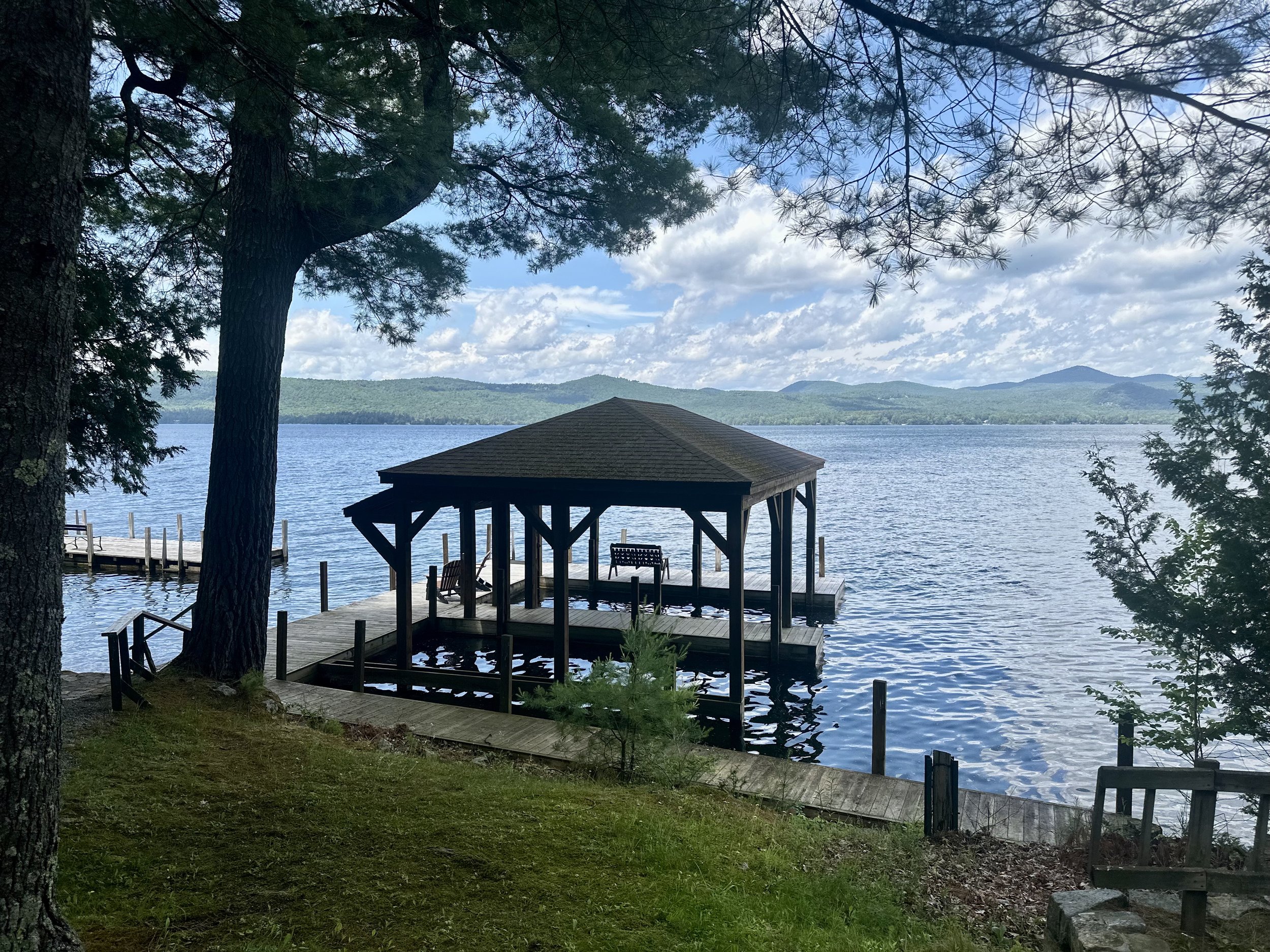 A lakeside scene with a wooden dock and gazebo, surrounded by trees, overlooking a body of water with distant hills under a partly cloudy sky.
