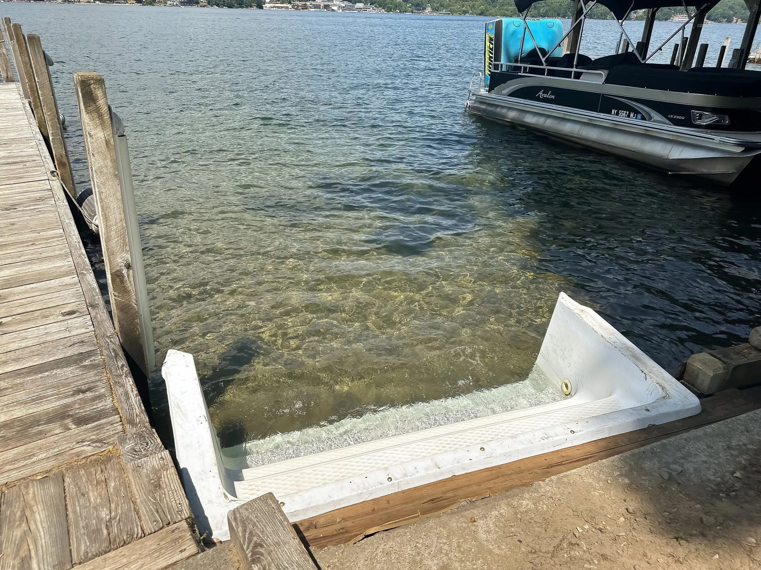 A boat dock at a lake with a black pontoon boat docked to the right and a wooden dock on the left. The water is clear and calm, with a shoreline and trees in the background.