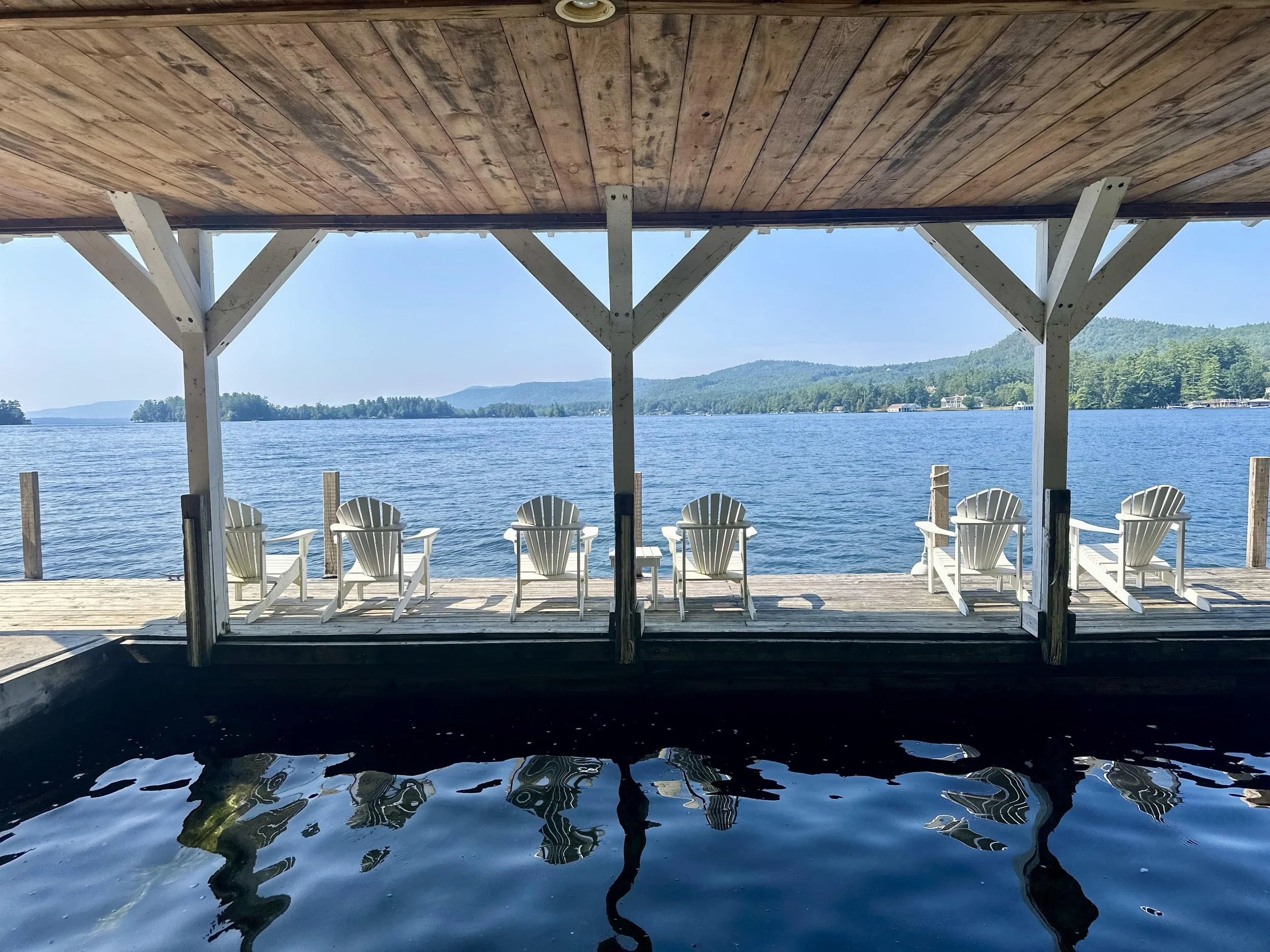 View of a lakeside porch with white Adirondack chairs, wooden ceiling, and support beams, overlooking a calm lake with distant trees and mountains under a clear blue sky.