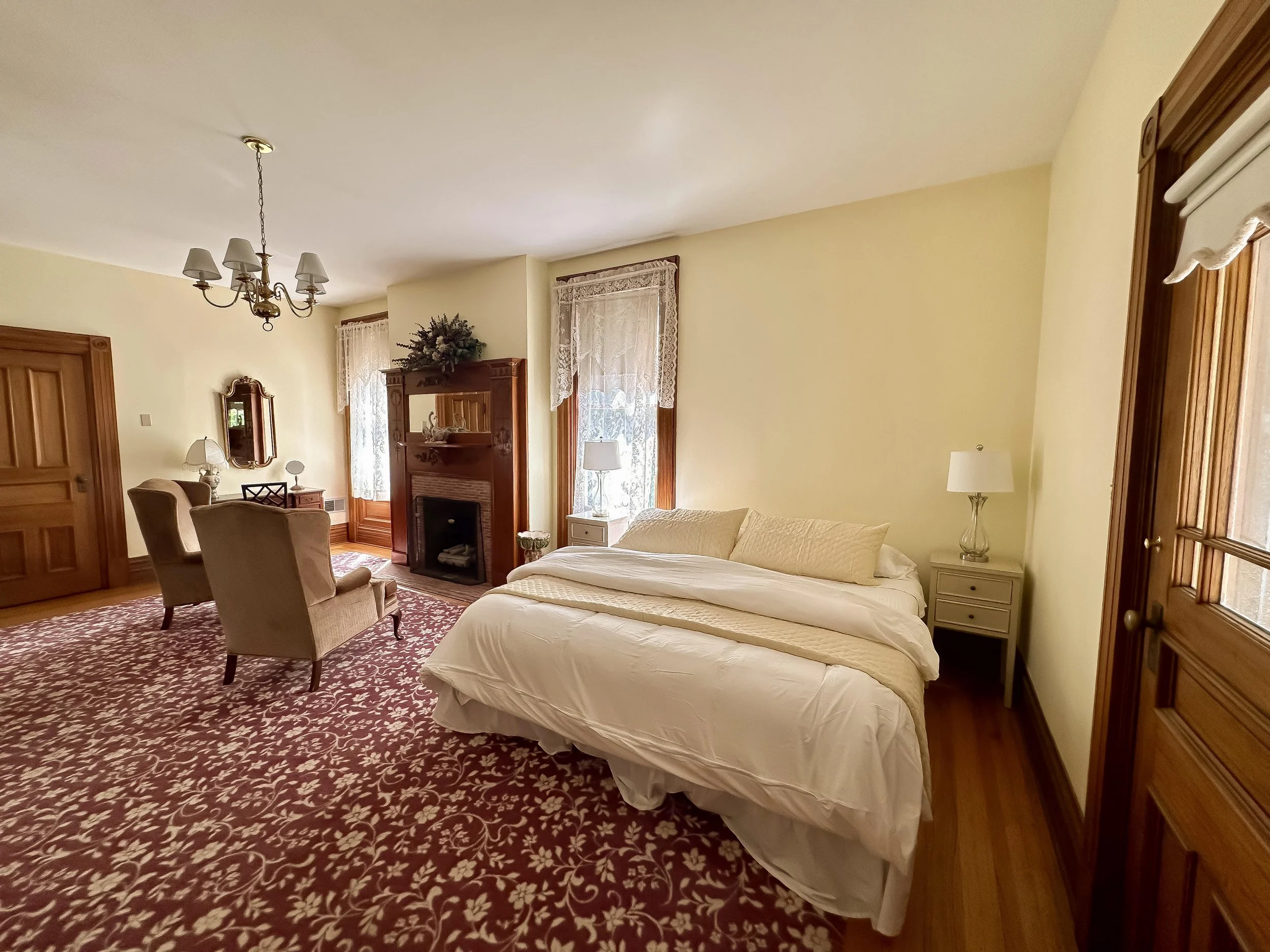 A cozy living room with beige walls, large windows with lace curtains, and wooden trim. The room contains a fireplace, a chandelier, a sofa, armchairs, and side tables with lamps, all on a patterned red and white carpet.