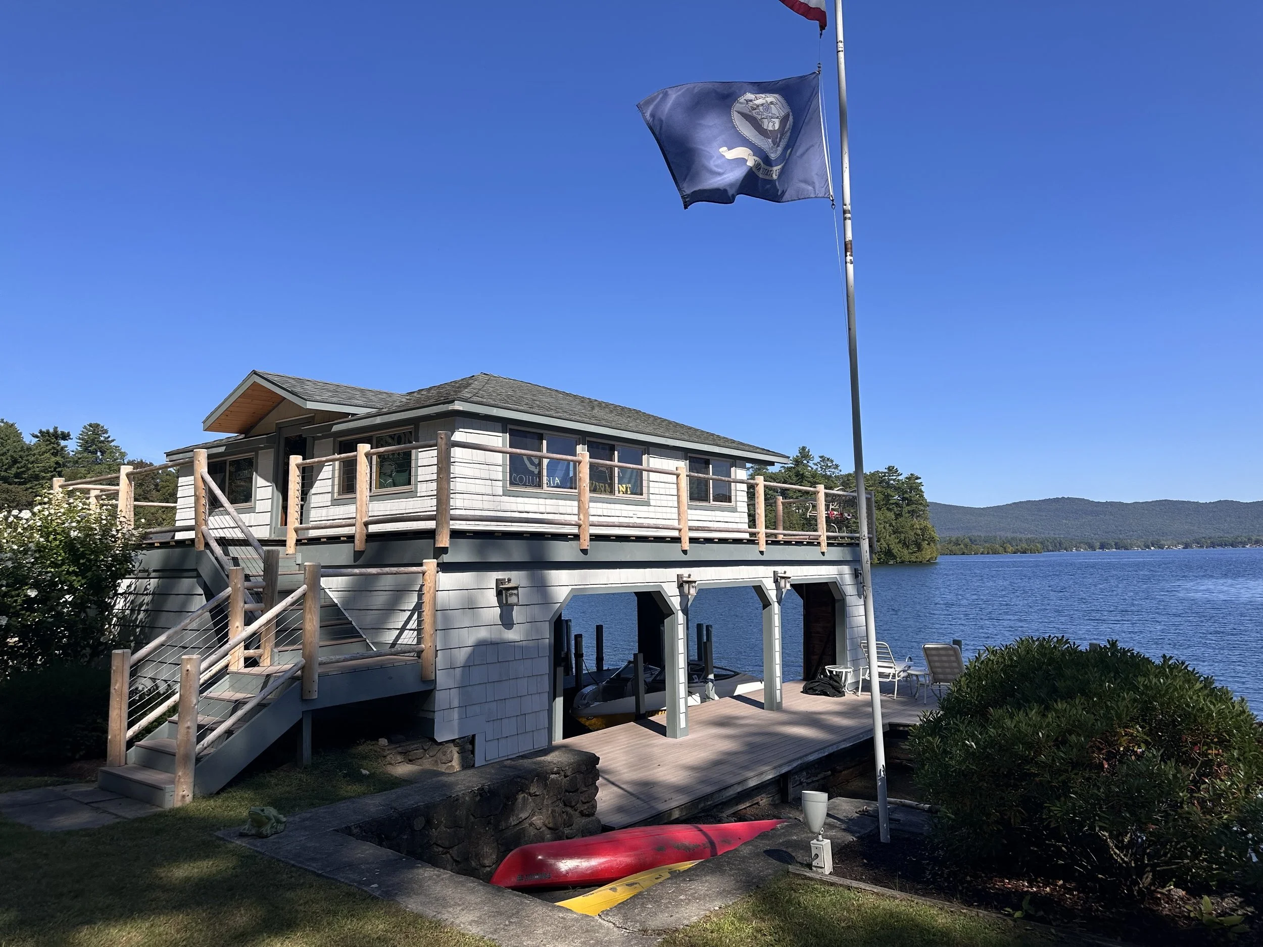 A lakeside house with a wooden deck and stairs, surrounded by greenery, with a flagpole flying a flag and a view of the lake and mountains in the background under a blue sky.