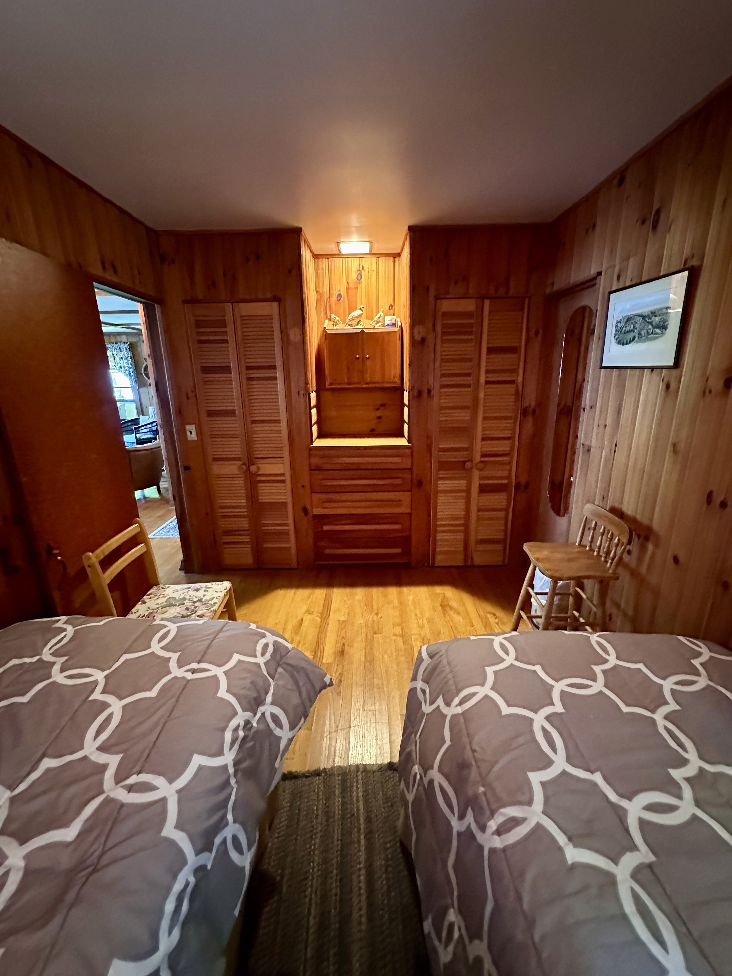 Bedroom with wood-paneled walls and two twin beds with matching patterned bedspreads, small chairs, and closet doors. A hallway leads to a room with a window and seating.